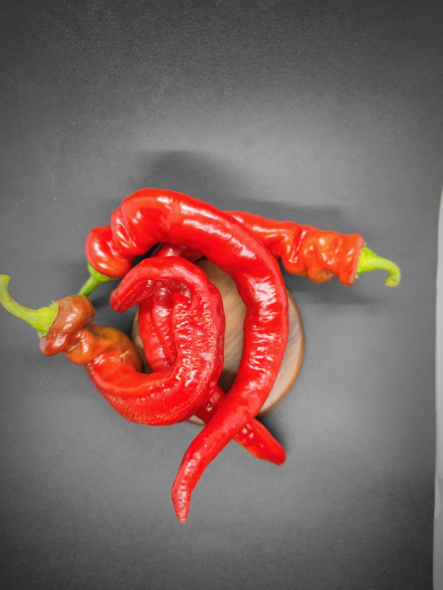 Three long, red Hangjiao 8 Total Eclipse (HJ8) chili peppers from PepperMerchant.net, each with a green stem, are arranged in a small wooden bowl against a dark background. The vibrant peppers, grown from space-grown seeds, have a slightly twisted and curved shape, exhibiting a fresh and glossy texture.