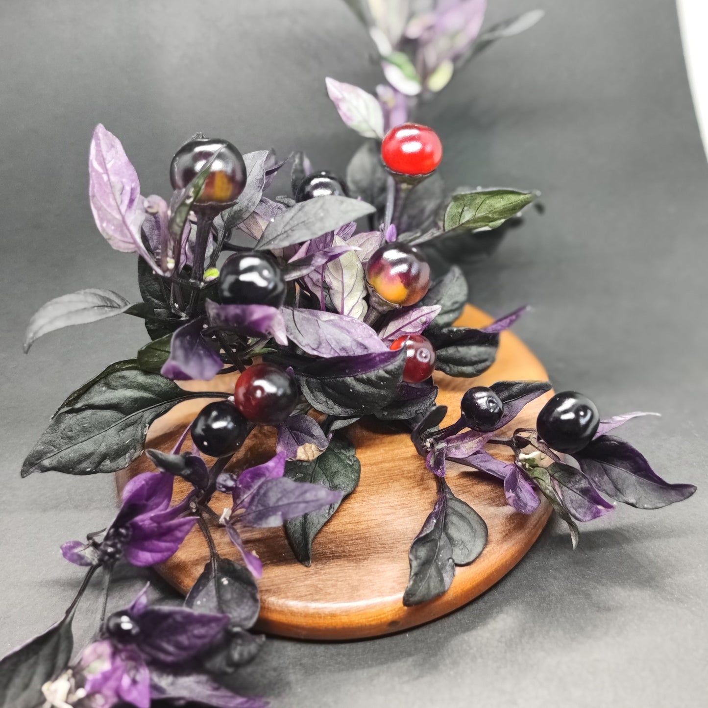 The Daybreaker by PepperMerchant.net features a branch with purple and green leaves plus glossy red, black, and dark purple berries—striped like pepper pods—displayed on a round wooden board against a dark background.