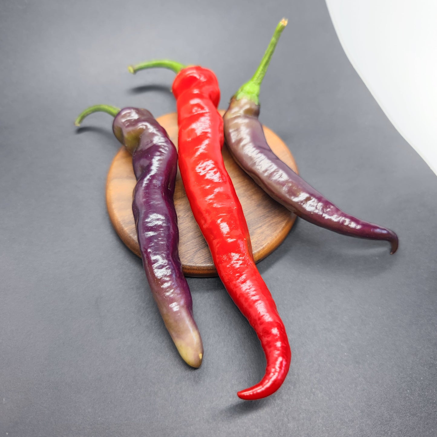 Three long Tiger-less Jalapeno peppers from PepperMerchant.net—two color-shifting purple and one red—are arranged on a small round wooden board against a dark gray background.