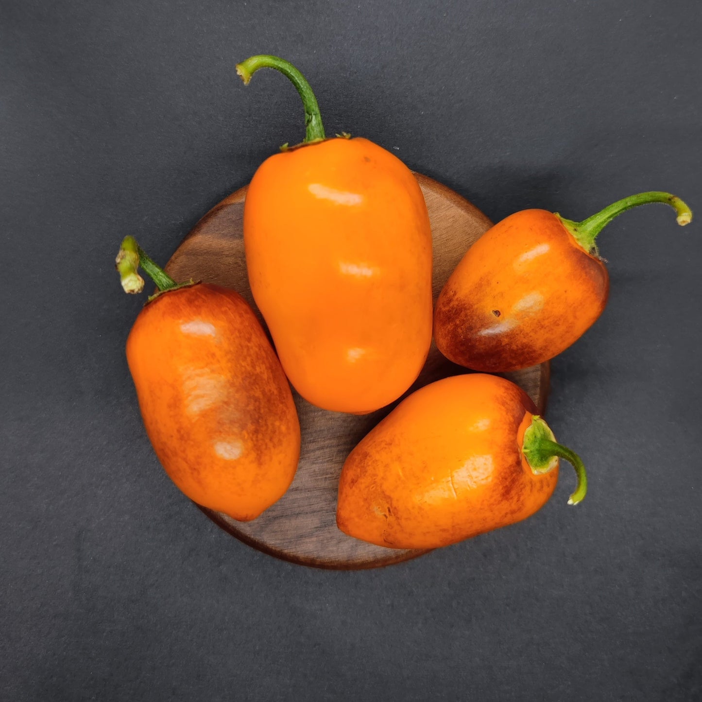 Four Desert Peach Rocoto peppers from PepperMerchant.net, some with brown blemishes, are arranged on a round wooden plate against a dark background—ideal as versatile cooking peppers or for those seeking moderate heat varieties.
