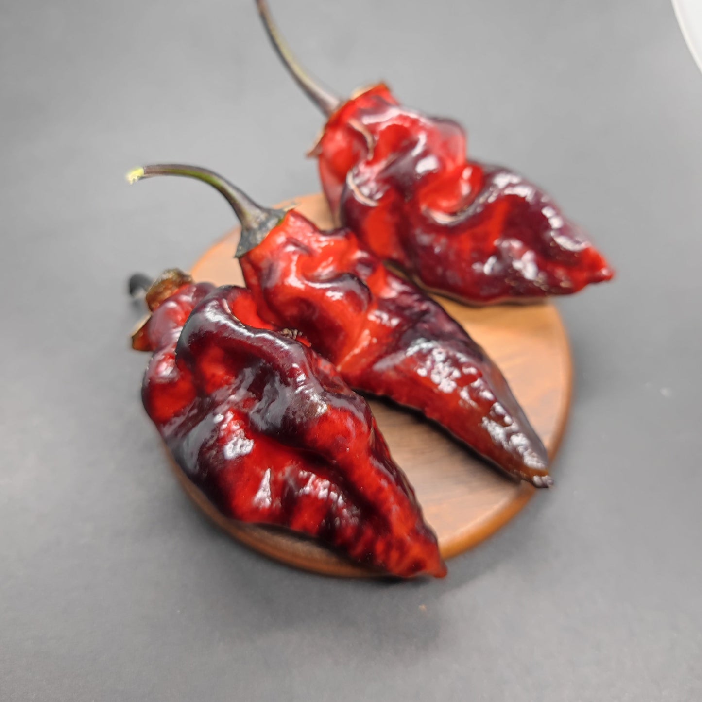 Three dark red, wrinkled BBG7 Alchemy - Dark Red Pheno peppers from PepperMerchant.net with green stems rest on a small wooden surface against a dark background, highlighting their superhot intensity.
