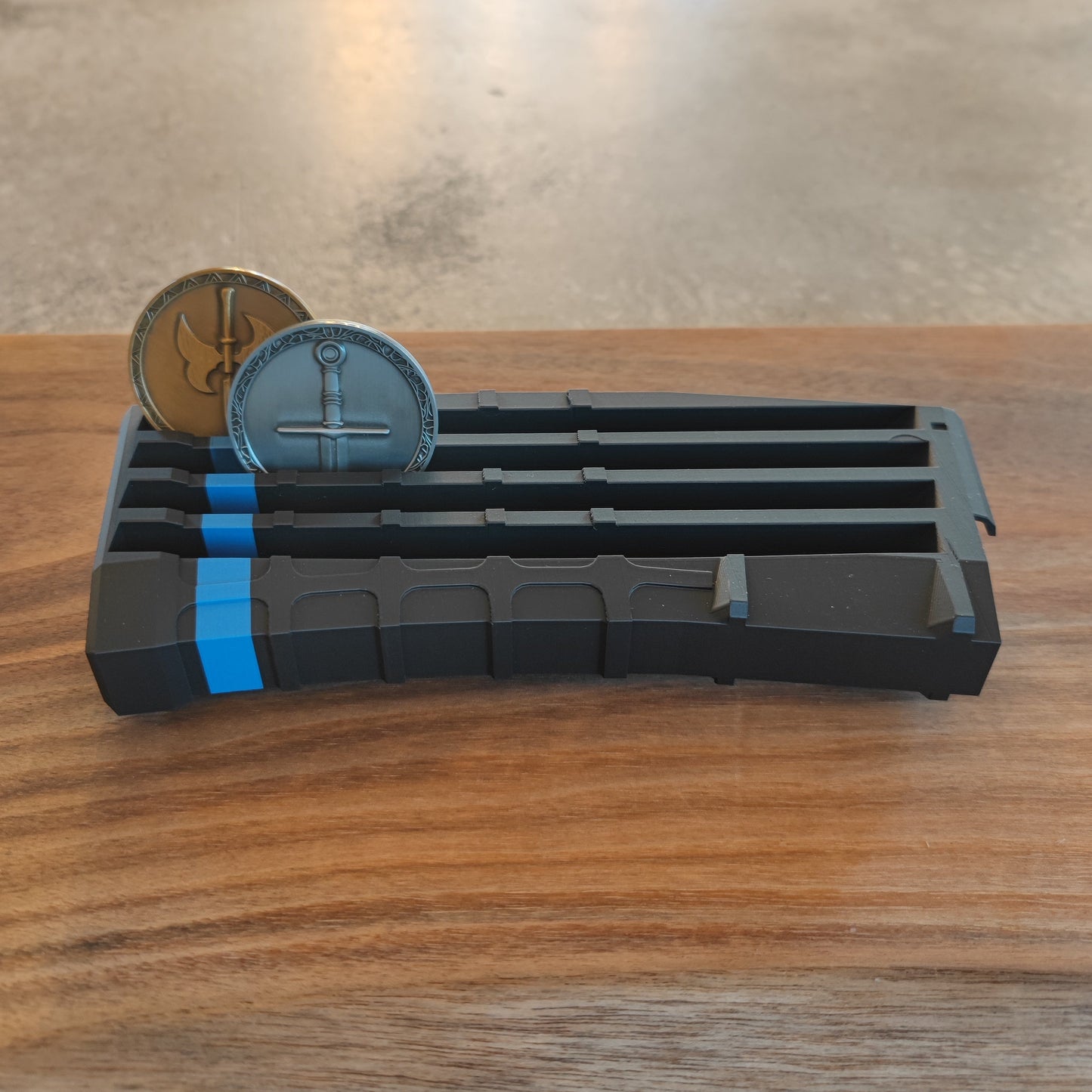 The GEN4 CoinMag - Blue Line by coin-mag.net is a black, rectangular holder with blue accents that displays two round coins upright in separate slots. It’s shown on a wooden surface with a blurred gray background.