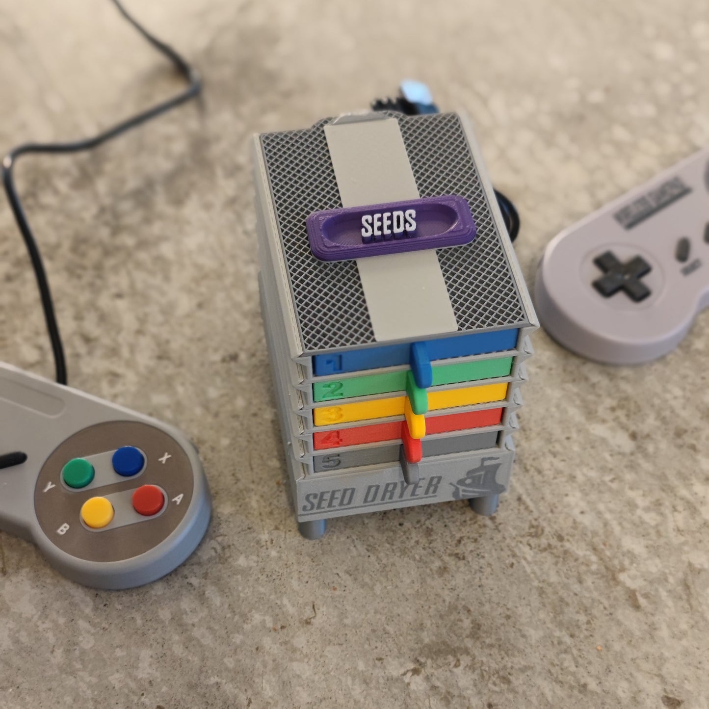 The PepperMerchant.net Seed Dryer - Super Classic Console is a small, gray, multi-level USB-powered seed dryer with colorful trays labeled 1-5, placed on concrete between two gray retro-style game controllers.