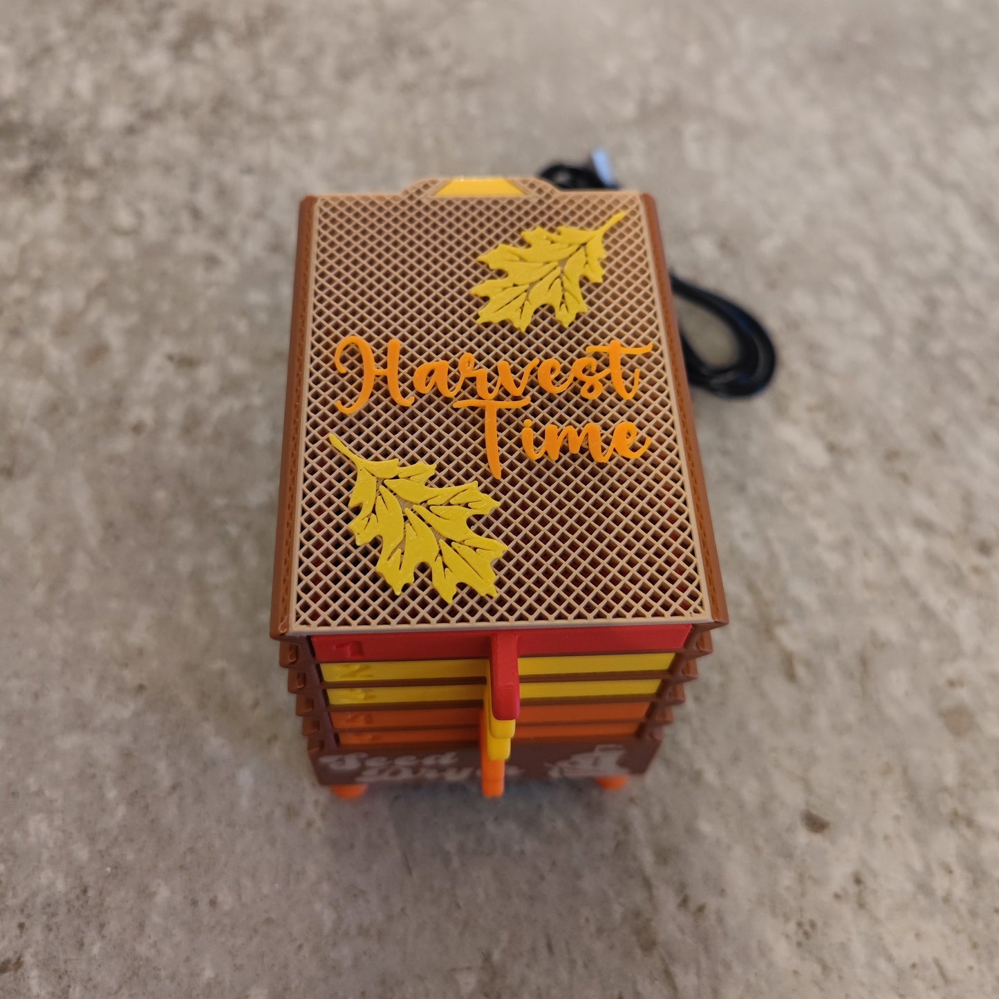 The Seed Dryer - Harvest Time by PepperMerchant.net is a small decorative box with a mesh-patterned lid, yellow leaves, and "Harvest Time" on top—ideal for seed saving or gentle drying—shown on a textured beige surface.