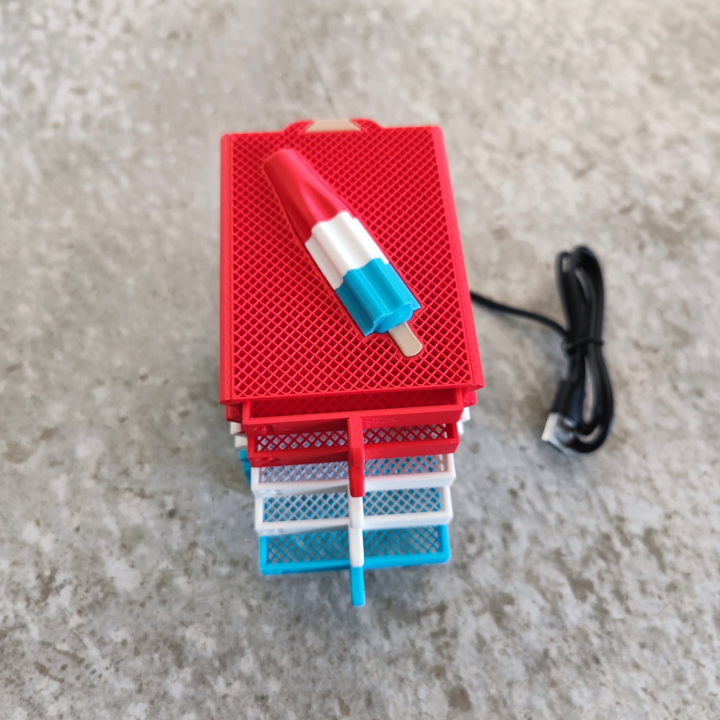A stack of colorful mesh cards topped with a Rocket Pop-shaped object sits next to a black USB cable on a textured gray surface—the Seed Dryer - Rocket Pop by PepperMerchant.net, an innovative USB-powered device designed for modern seed saving technology.