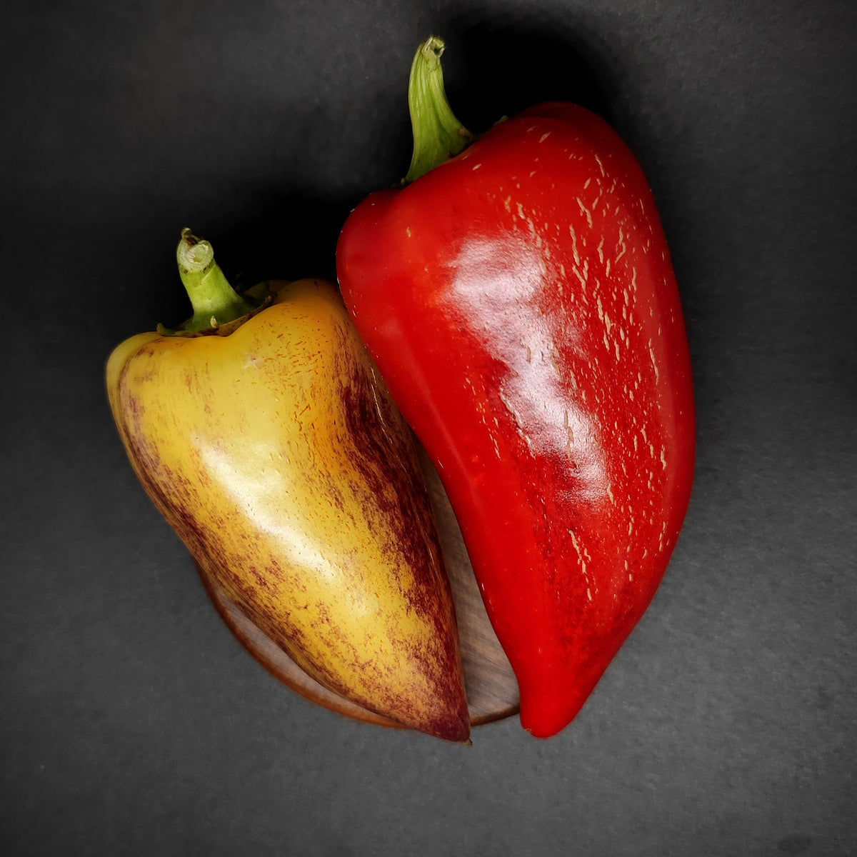 A Violet Sparkle thick-walled yellow pepper with dark streaks and a bright red cooking pepper rest on a small circular wooden base.