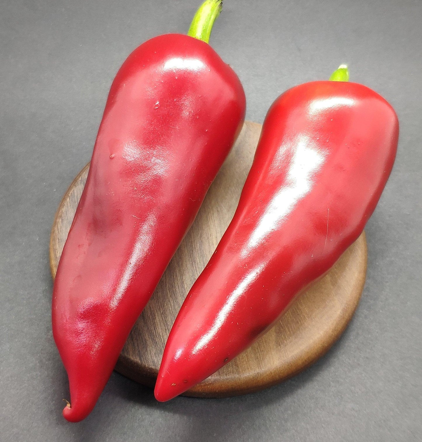 Two large, glossy red Espelette Peppers from PepperMerchant.net with green stems rest on a round, wooden surface against a dark background.