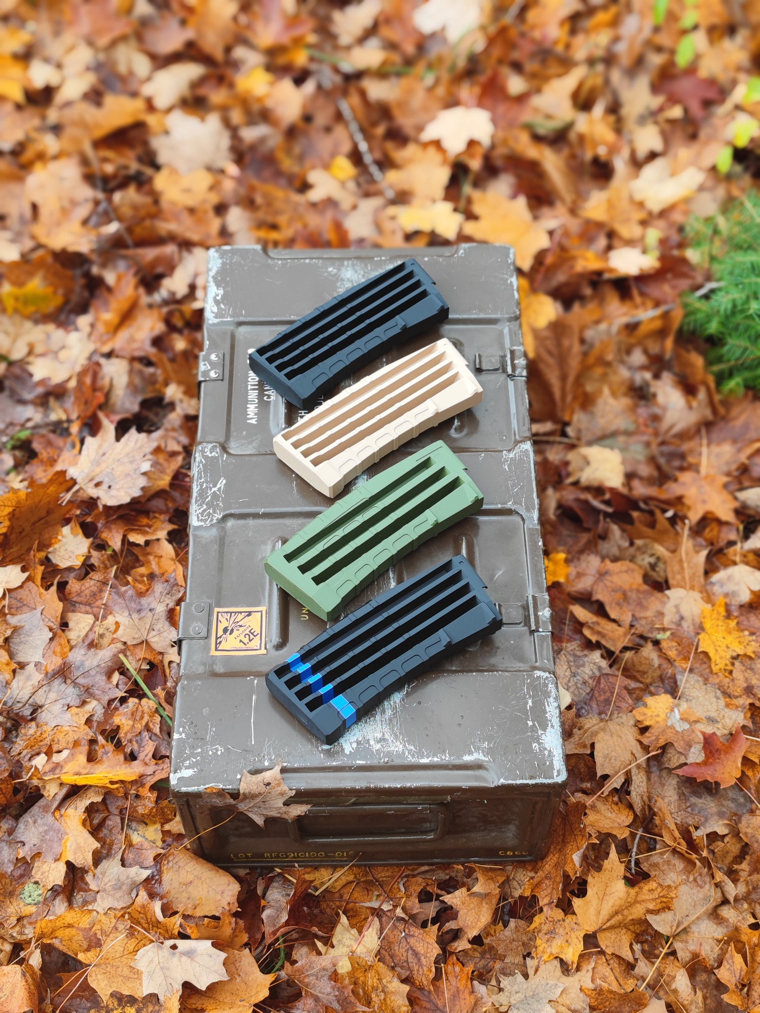 Four rifle handguards—black, tan, green, and blue-tipped—rest on a weathered ammo box surrounded by autumn leaves.