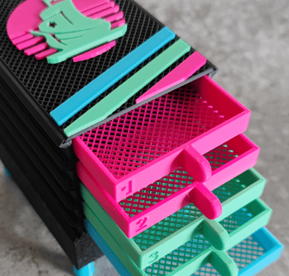 A colorful set of plastic drawers with turquoise, pink, and black compartments, labeled with numbers and symbols. The drawers are partially open, showing a mesh-like design. They are stacked on a grey, textured surface.