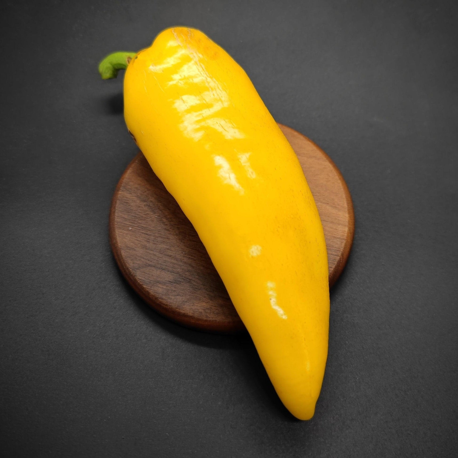 A single XL Lemon Spice Jalapeno from PepperMerchant.net is displayed on a round wooden coaster against a dark background. The pepper is glossy and elongated, with a slight curve and a green stem at the top. The lighting highlights its vibrant color and smooth texture.