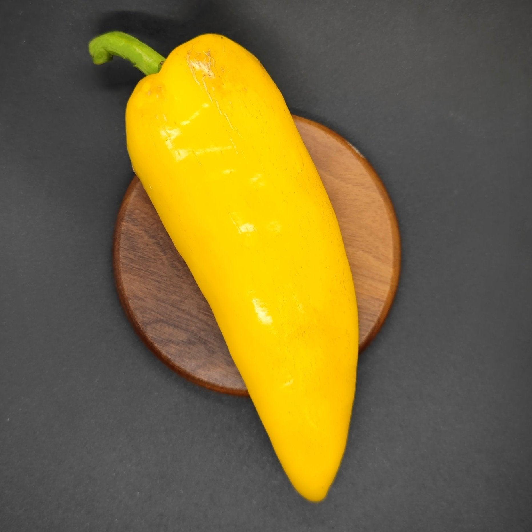 An XL Lemon Spice Jalapeno from PepperMerchant.net with a green stem is placed on a small, round wooden plate against a dark background.