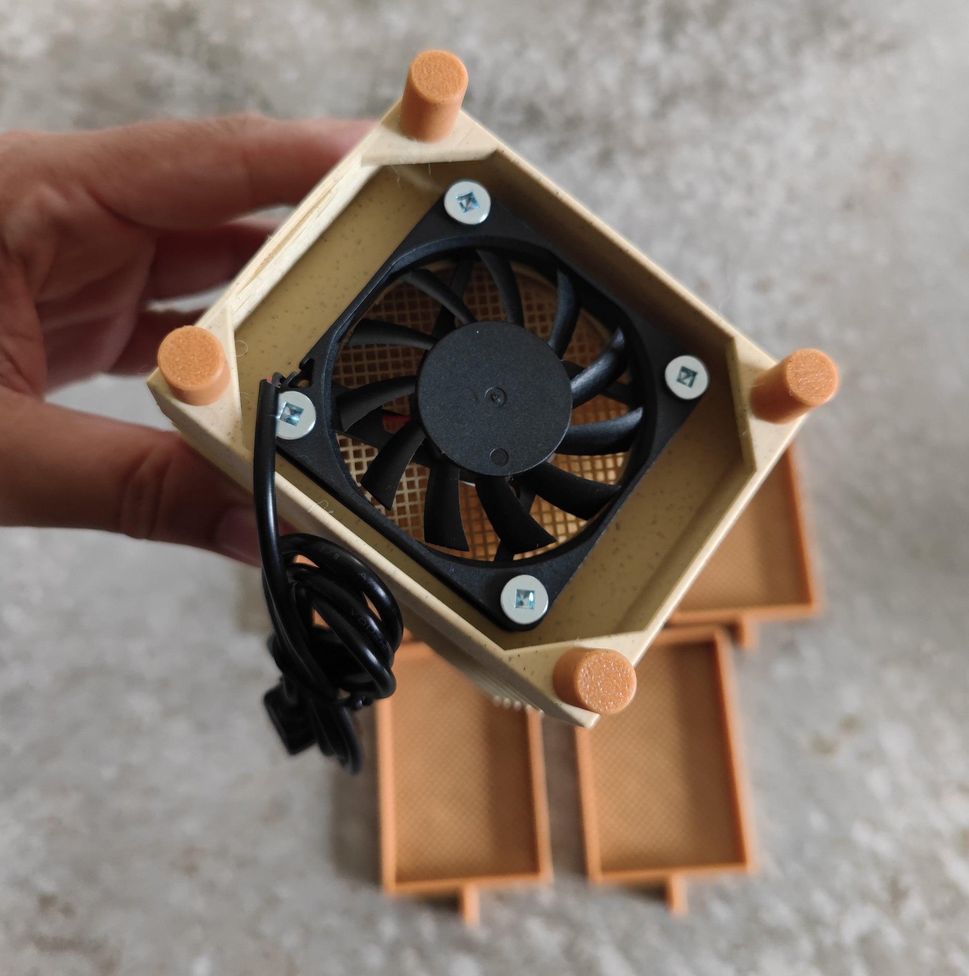 A person holding a tan, rectangular Seed Dryer - Pumpkin Spicy *Limited Edition only 8 Made* from PepperMerchant.net, featuring a small black cooling fan and coiled cable. The USB-powered device has orange rubber feet on each corner, with two similar dryers blurred in the background on a concrete surface.