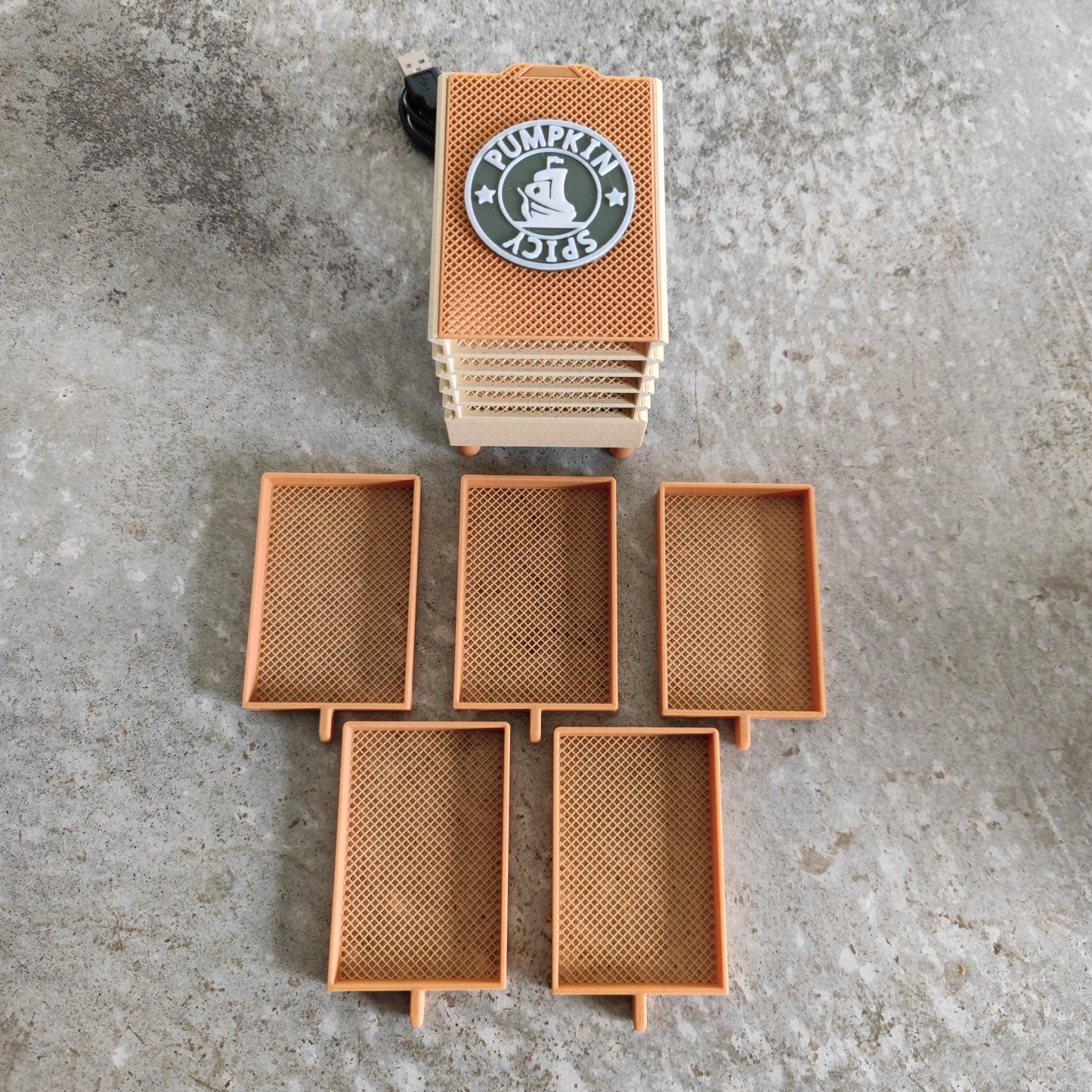 A beige Seed Dryer - Pumpkin Spicy *Limited Edition only 8 Made* from PepperMerchant.net sits on a concrete floor. The top lid displays a pumpkin logo with the words "Pumpkin Spicy." Below the dehydrator, five removable drying trays are spread out, showcasing their rectangular shape and mesh design, exemplifying advanced seed-saving technology from PepperMerchant.net.