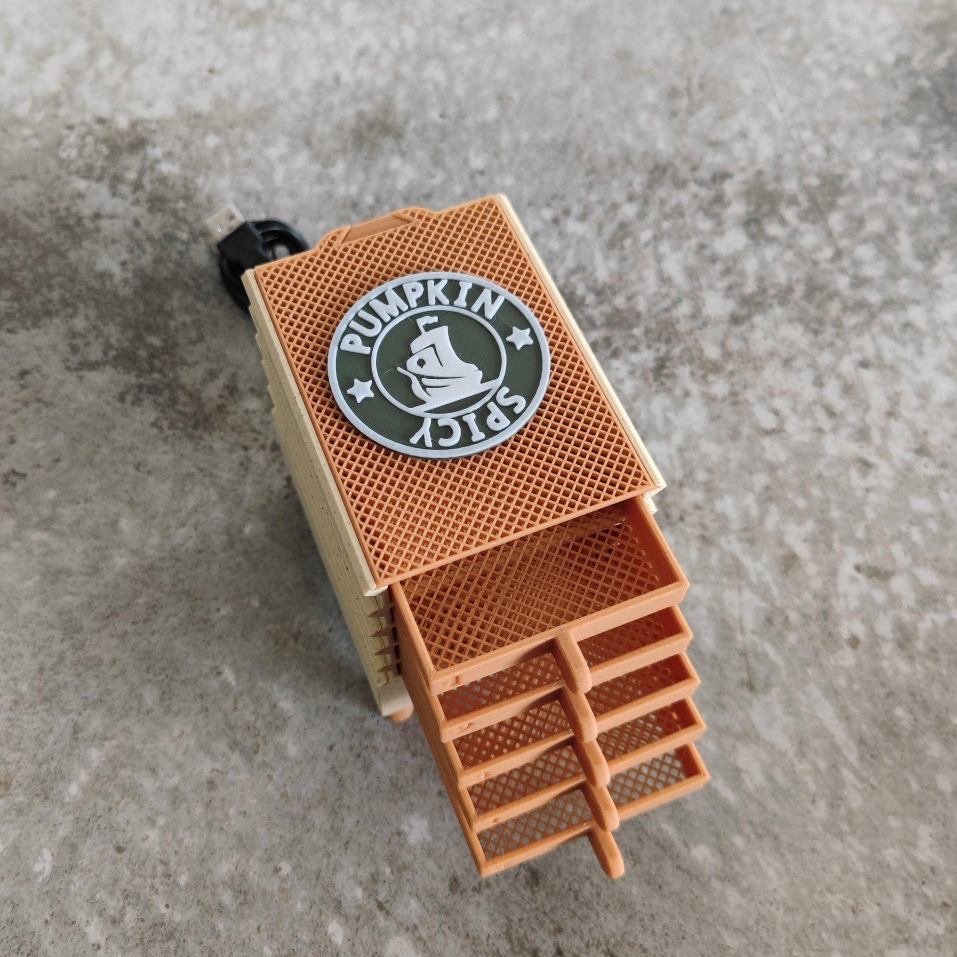 A tan and orange mesh box labeled "Seed Dryer - Pumpkin Spicy *Limited Edition only 8 Made*" from PepperMerchant.net, featuring a circular logo, sits on a gray surface. The box has multiple stackable layers suggesting specialized storage, and a black cord extends from one end, indicating it is likely an electronic device.