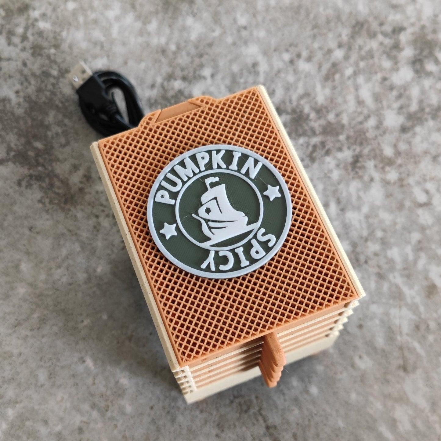 A limited edition Seed Dryer - Pumpkin Spicy from PepperMerchant.net, only 8 made, sits on a grey surface. The portable device features a textured brown and beige casing with a circular emblem that reads "Pumpkin Spicy" alongside an image of a coffee cup. A black USB cable is attached to the top, showcasing the compact convenience of Pumpkin Spicy seed dryer technology.