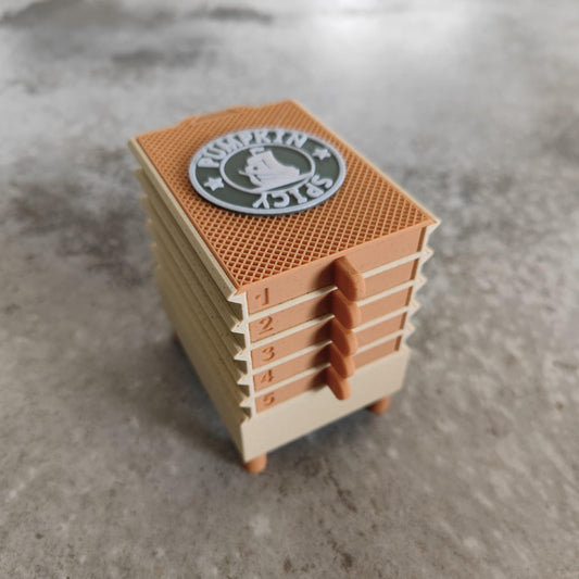 This small, tan and orange stackable model resembles a chest of drawers and is labeled with numbers 1 through 5 on the side. The top features a circular logo with a bell emblem and text. Perfect for organizing seeds, it rests on a gray, textured surface—making it an ideal companion to your USB-powered Seed Dryer - Pumpkin Spicy *Limited Edition only 8 Made* from PepperMerchant.net.