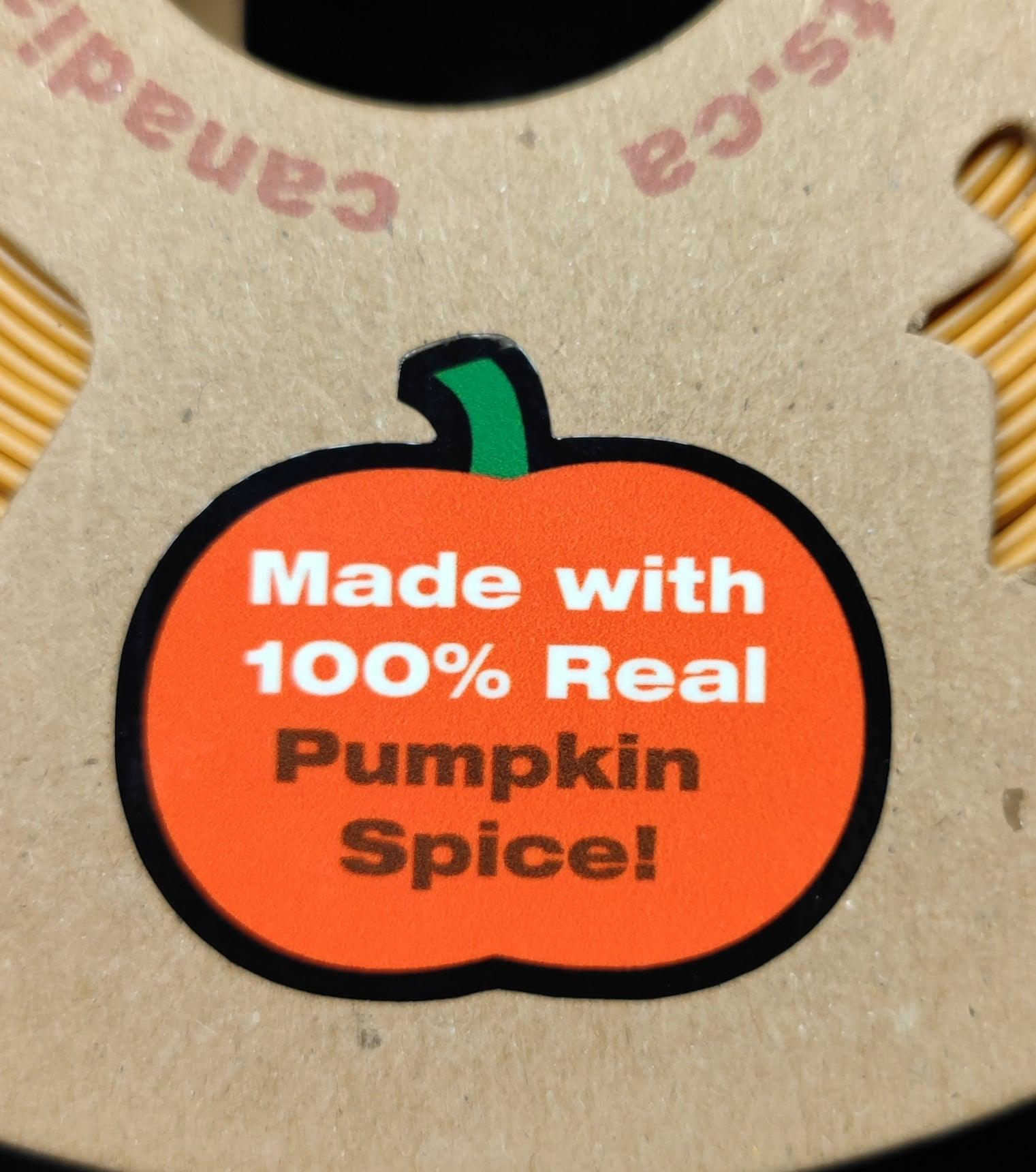 A cardboard surface features a pumpkin-shaped sticker with the text "Made with 100% Real Pumpkin Spice!" The pumpkin image has a green stem, and the text is in white and orange. Part of the surrounding text on the cardboard is partially visible at the top, celebrating PepperMerchant.net's Seed Dryer - Pumpkin Spicy *Limited Edition only 8 Made* seed saving technology.