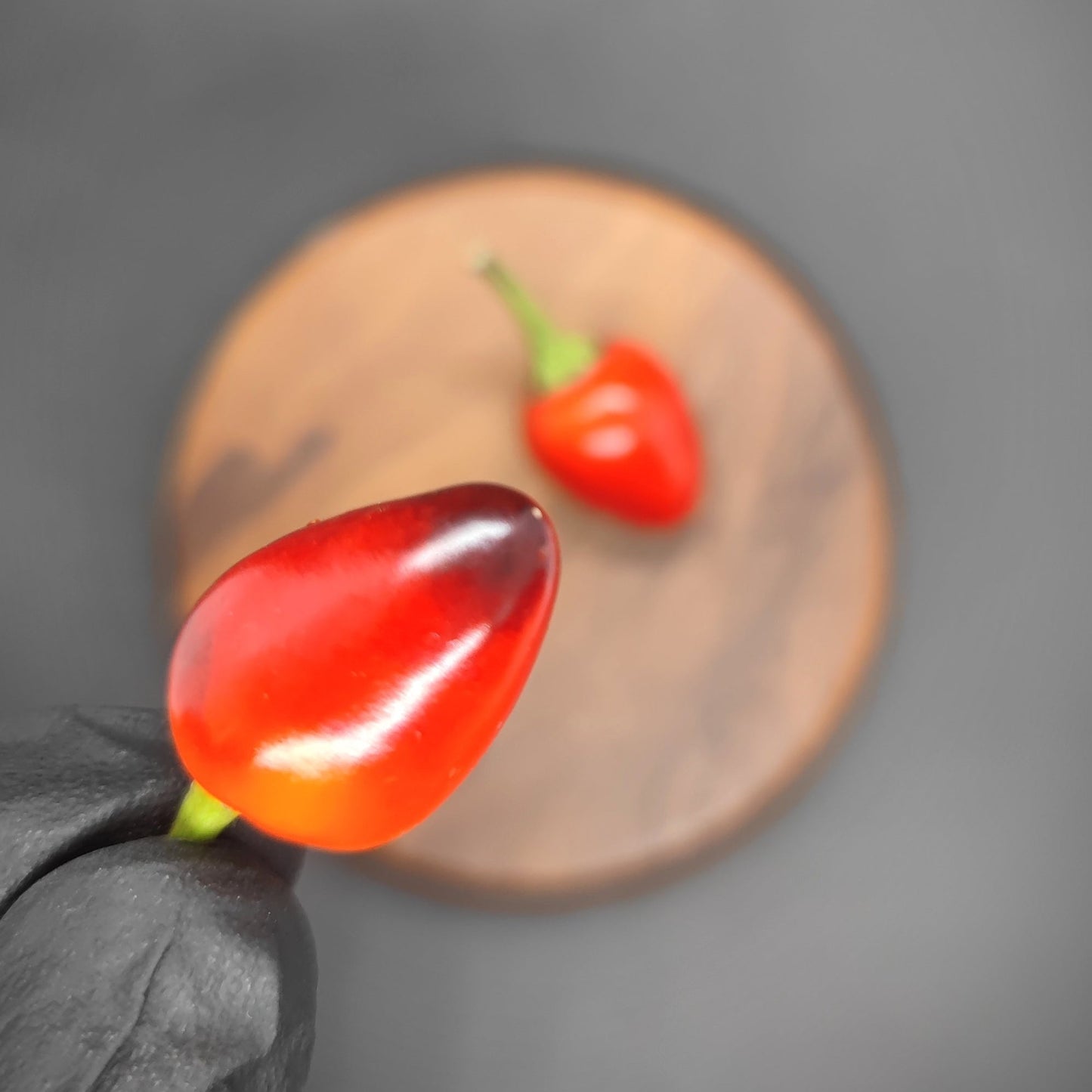 A gloved hand holds a small, shiny Midwest Sunset pepper with a green stem against a blurred background. Another similar Midwest Sunset pepper rests on a wooden round board in the backdrop. Both peppers are vibrant and appear fresh, reminiscent of the radiant hues seen during a Midwest Sunrise.