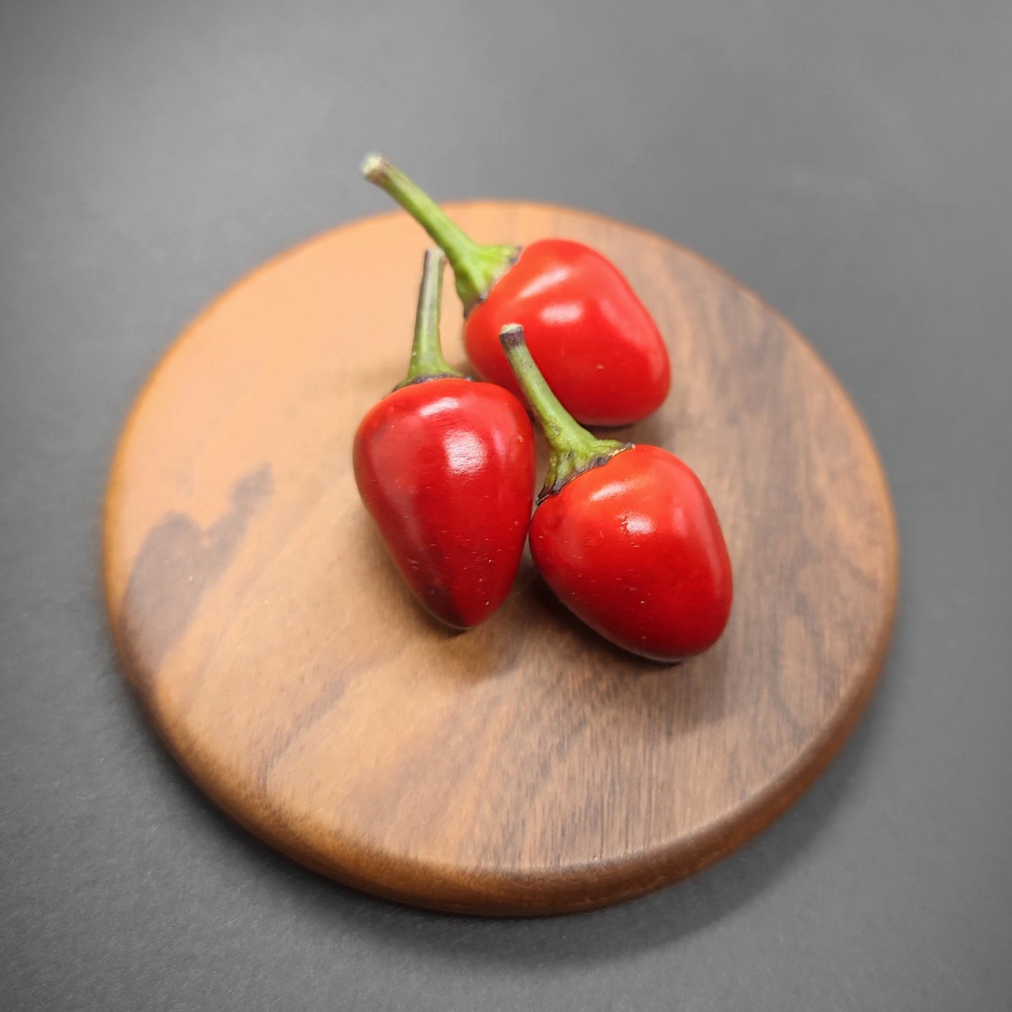 Three red chili peppers with green stems rest on a round wooden coaster, reminiscent of Midwest Sunset by PepperMerchant.net, against a dark, softly lit background.