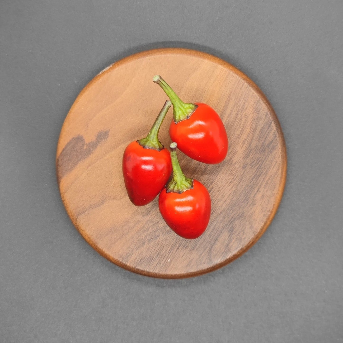 A trio of small, red chilis with green stems are artfully arranged on a round wooden board. The board is set against a Midwest Sunset (Red Midwest Midnight) backdrop from PepperMerchant.net, creating a striking contrast that accentuates the vibrant colors of the chilis.