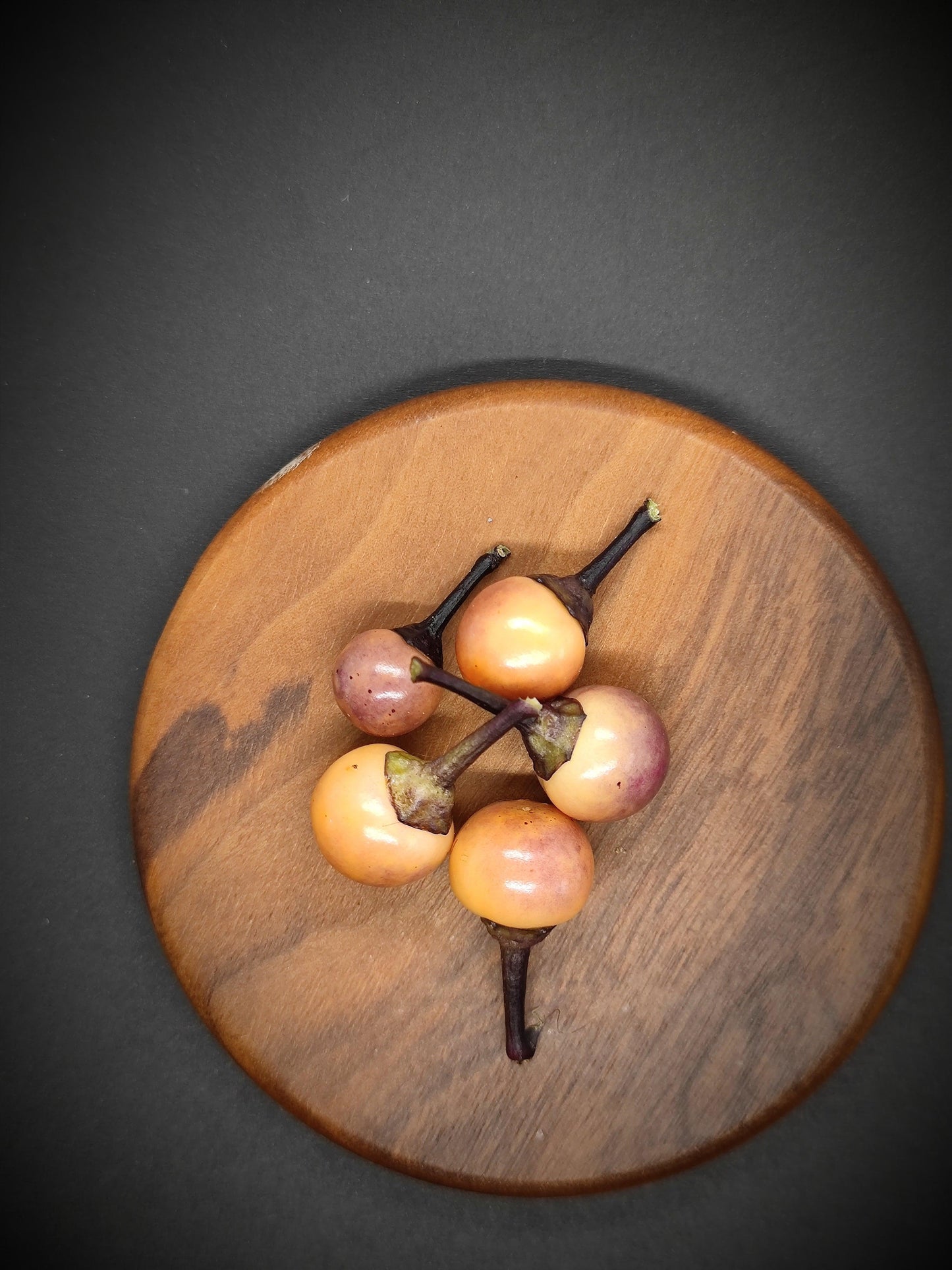 A wooden plate with five small, round, peach-colored tropical fruits with dark stems, arranged neatly in a cluster. The plate rests on a surface reminiscent of Midwest Midnight from PepperMerchant.net, creating a contrast that highlights the natural colors and textures of the fruits and wood.