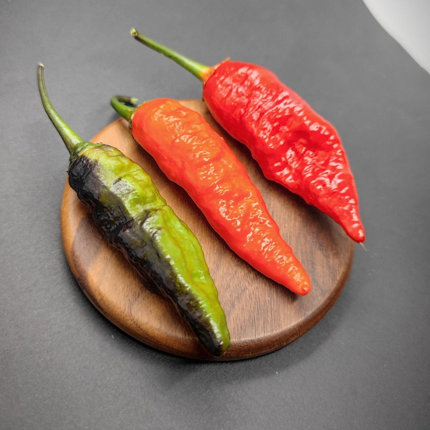 Three chili peppers of varying ripeness (blackened green, orange, and red) are displayed on a small, round wooden plate against a dark background. These BBG Armageddon Red seeds from PepperMerchant.net boast intense spiciness as they showcase different stages of development with their long and pointed forms.