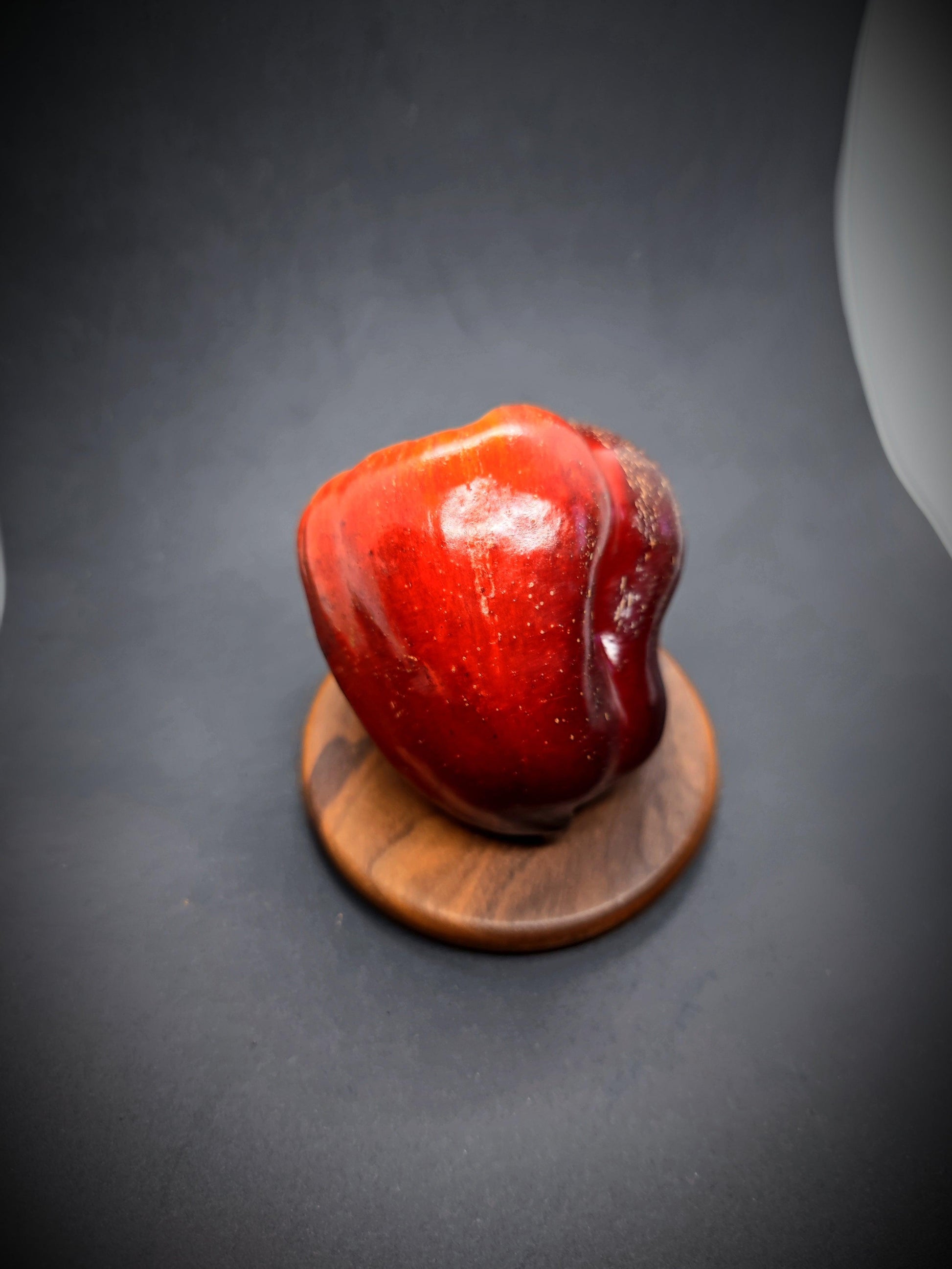 A single, shiny red Iko Iko sweet pepper from PepperMerchant.net stands on a round wooden disk, set against a dark background. The pepper's vibrant color contrasts sharply with the muted surroundings, making it the focal point of the image.