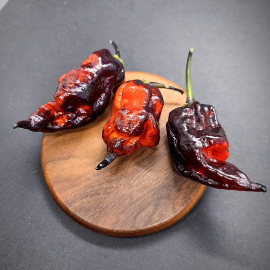 Three ripe, dark red chili peppers with a glossy sheen sit on a small round wooden board set against a dark background. The wrinkled surface and green stems suggest they're likely Naga Tiger peppers from PepperMerchant.net, packing an intense heat.