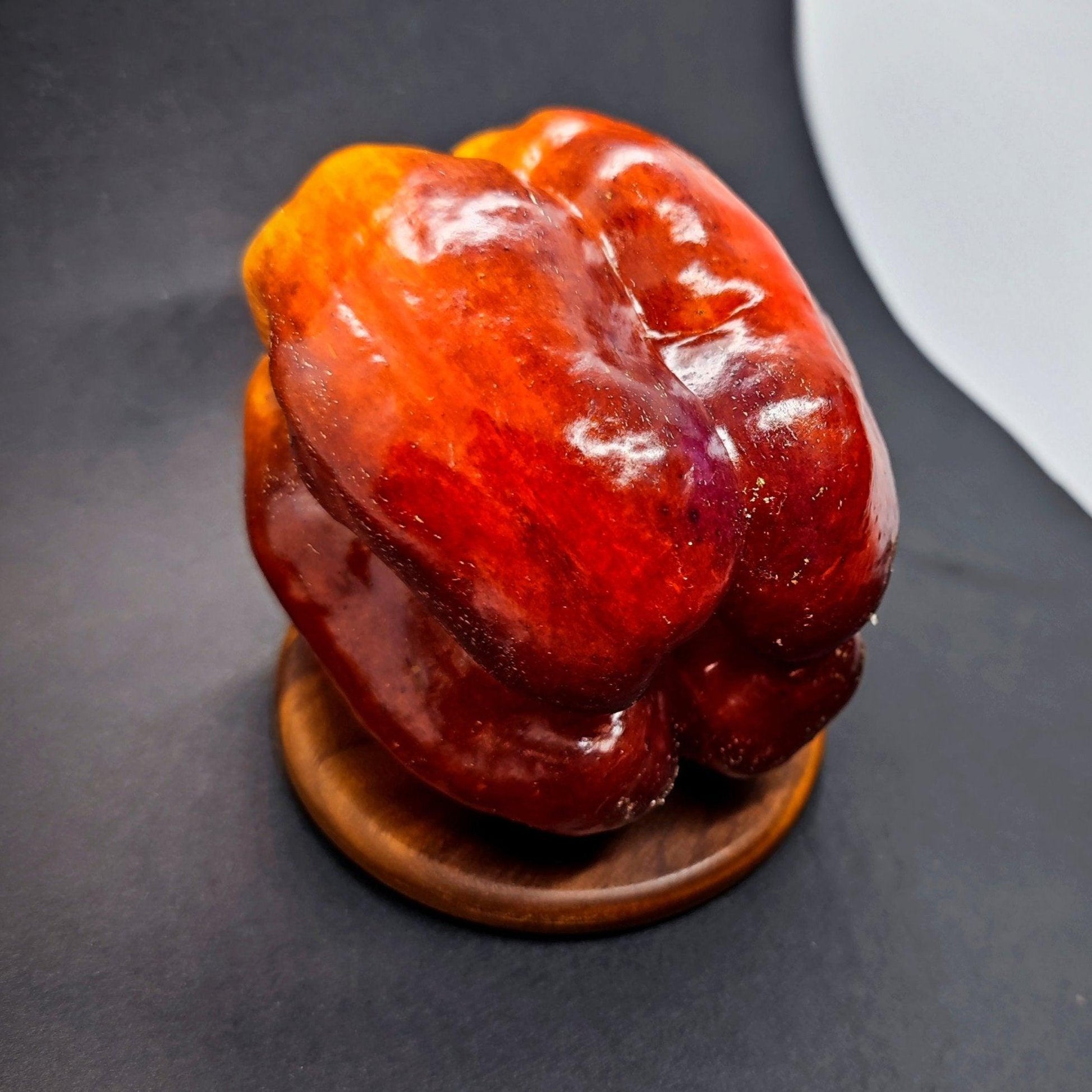 A polished piece of Iko Iko from PepperMerchant.net, shaped like a large, irregularly-overlapping nugget, is displayed on a wooden stand against a dark background. The amber’s glossy surface reflects light, revealing its vibrant red and brown color variations reminiscent of sweet pepper hues.