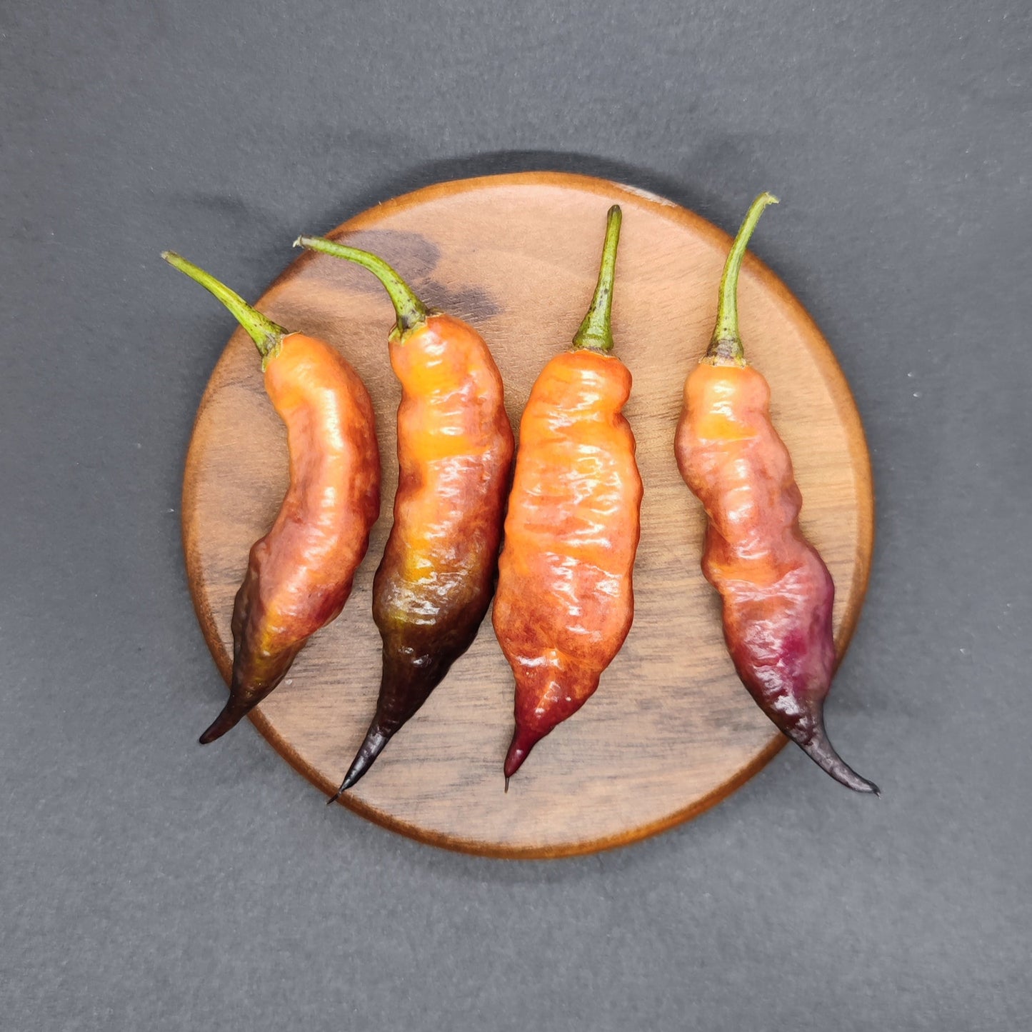 The "Pseudo Ganzo Pastel" from PepperMerchant.net features four vibrant chili peppers, including the unique Reaper Pink, with stunning gradient hues of red, orange, and purple arranged on a round wooden board against a dark background.