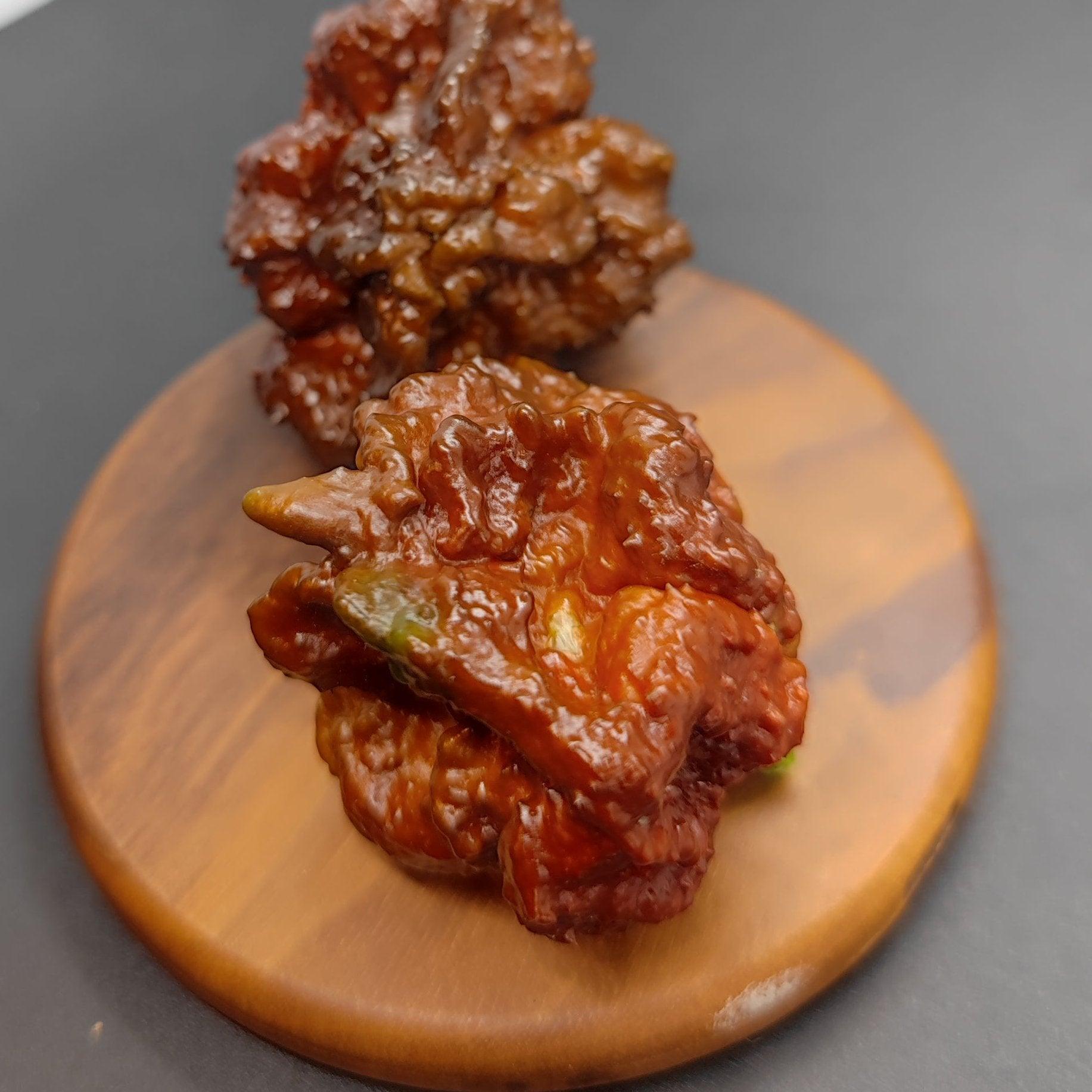 Two pieces of Korean fried chicken, coated in a sticky, reddish-brown sauce featuring a hint of Chocolate Brainstrain pepper from PepperMerchant.net, are placed on a small, round wooden board set against a contrasting black and white background. The chicken pieces have a crispy and textured appearance.