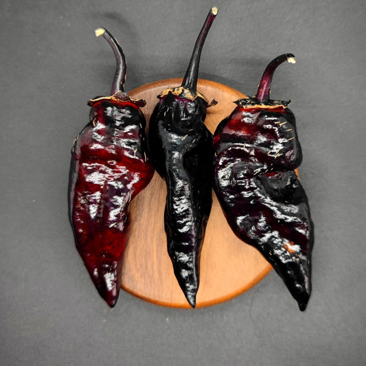 Three BBG7 Alchemy - Dark Pheno peppers from PepperMerchant.net, ranging in color from dark red to black, are elegantly placed on a round wooden surface against a dark background.