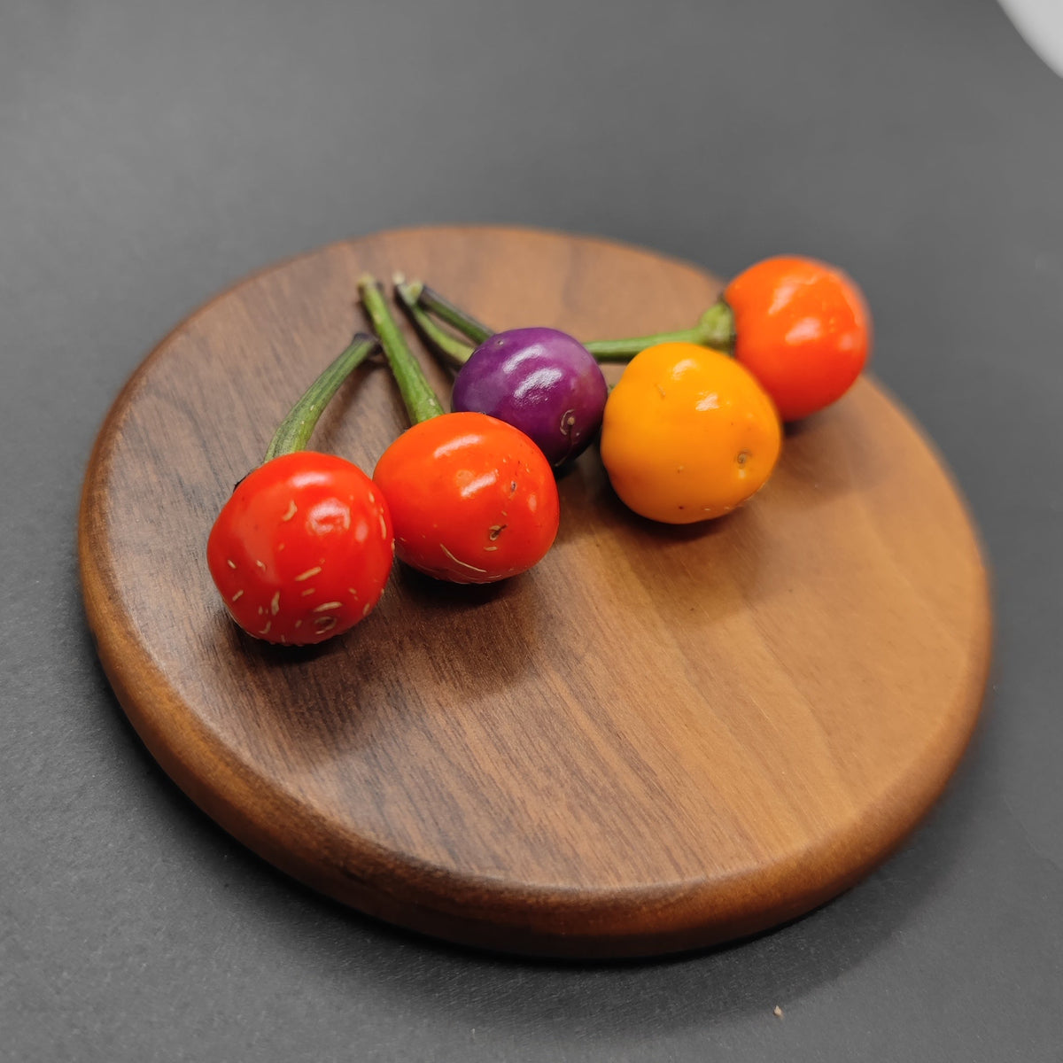 Five small, round Faddas AC101B peppers from PepperMerchant.net—resembling Italian peppers—display vivid shades of red, orange, yellow, and purple. Arranged with their stems clustered on a circular wooden board against a black background, they hint at their mild heat and vibrant character.