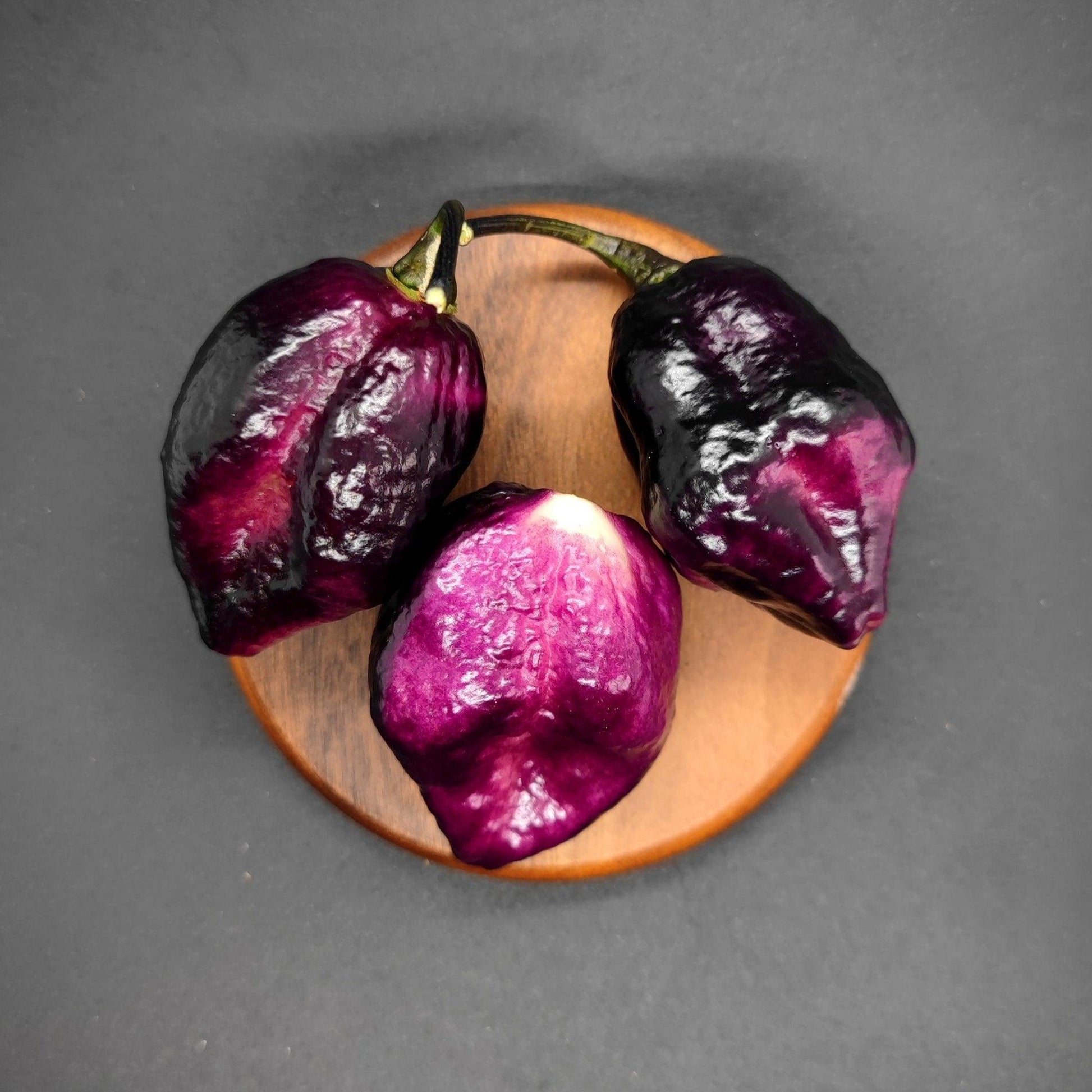 Three Phat Karen peppers from PepperMerchant.net, each with a glossy sheen and uniquely shaped, are arranged in a triangular formation on a round, wooden surface against a plain, dark background. The variations in shades of purple and black make these striking purple pods stand out.