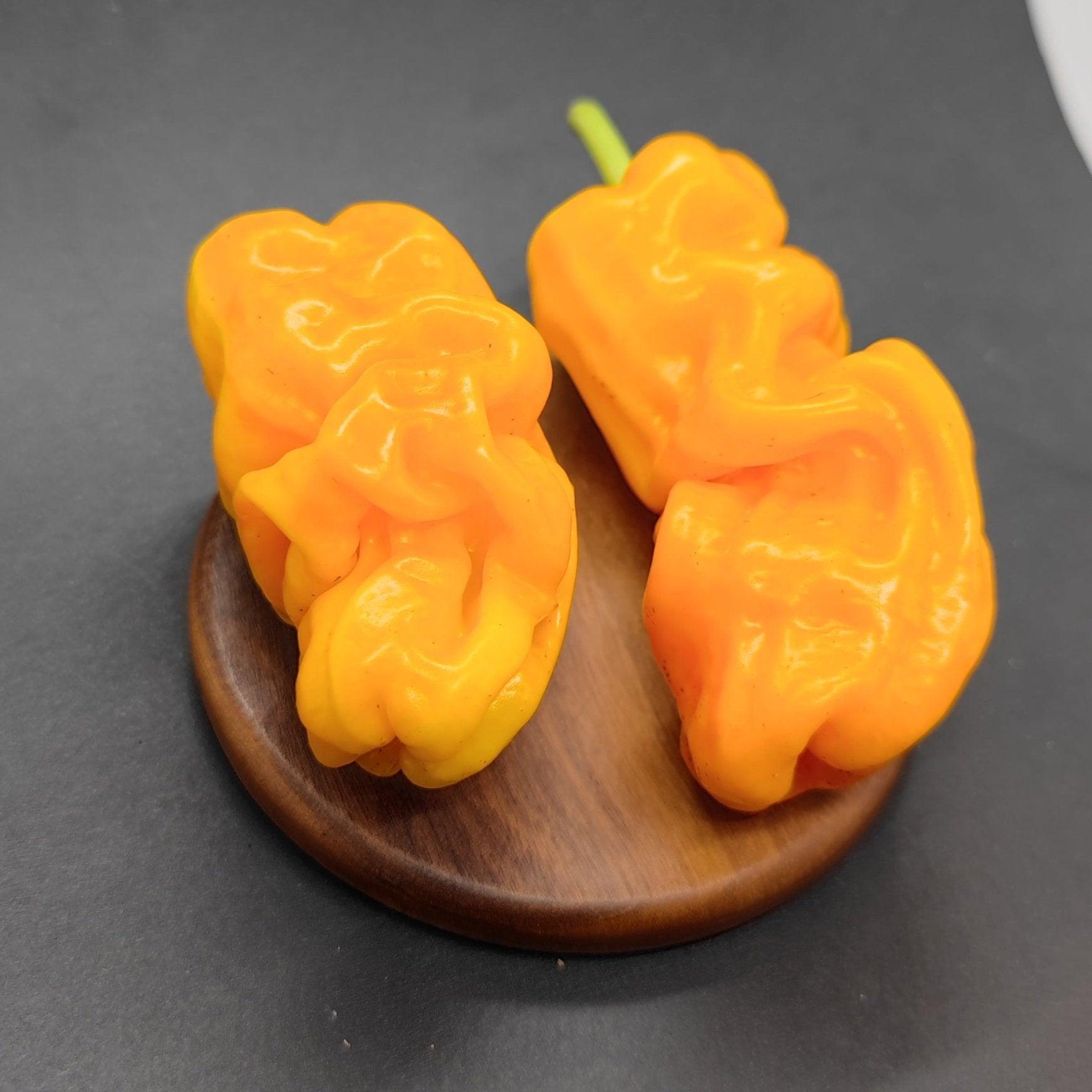 Two vibrant yellow-orange Dragons Toe peppers from PepperMerchant.net sit on a small, round wooden platform against a black background. The peppers have a glossy surface and irregular, bumpy shapes, making them perfect for adding an exotic twist to Asian dishes.