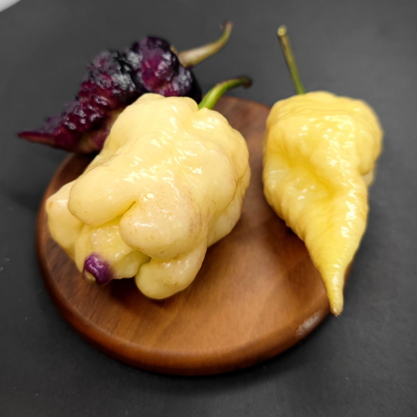 Three superhot peppers from PepperMerchant.net's Jays x Pink x Lemon Reaper collection, including a dark purple one similar to the Purple Haze variety and two pale yellow ones, are displayed on a small wooden round board against a dark background. The peppers boast glossy surfaces with unique, wrinkled textures.