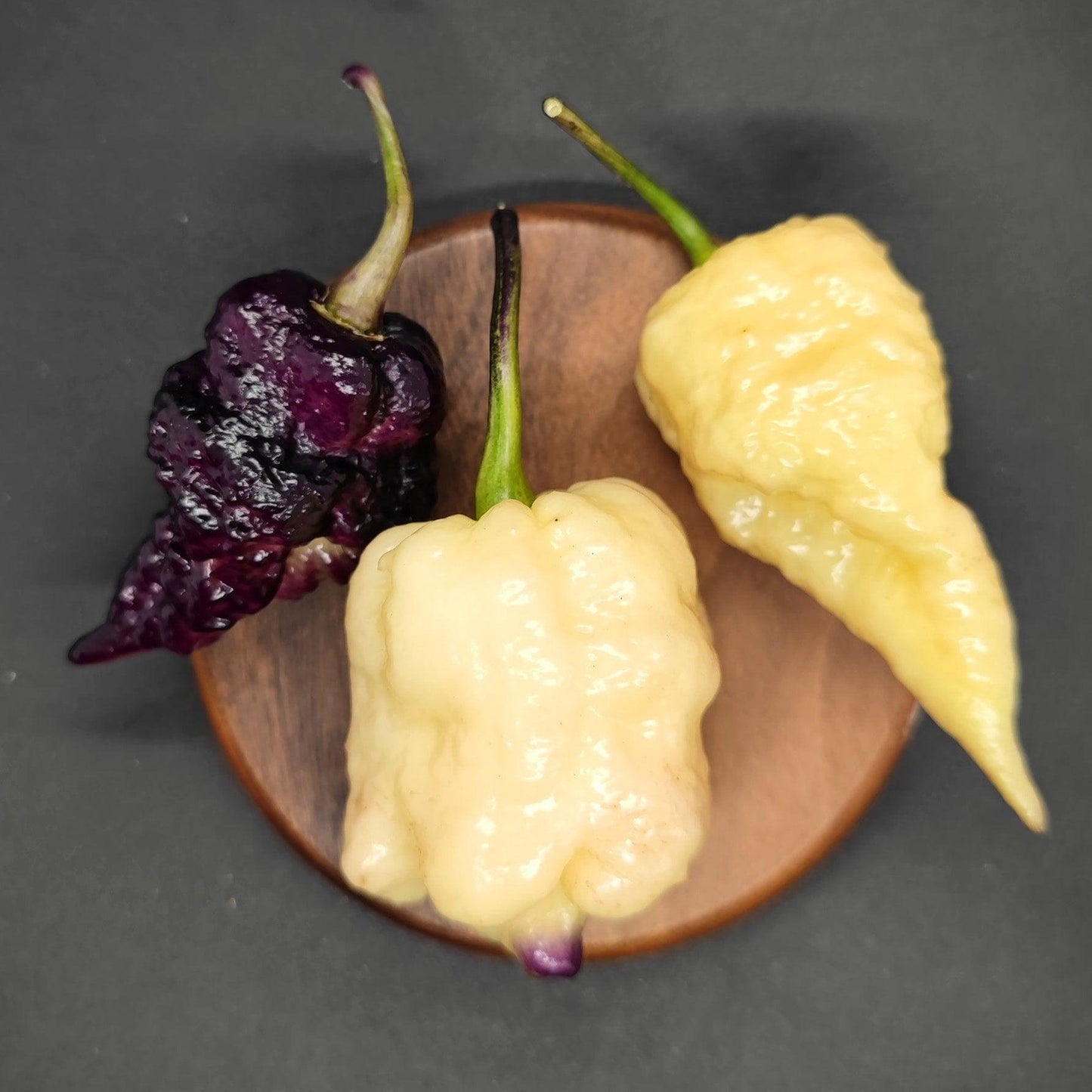 A wooden plate displays three unique varieties of the superhot Jays x Pink x Lemon Reaper peppers from PepperMerchant.net. On the left, there's a deep purple haze pepper; in the center, a pale yellow one with a bumpy texture; and on the right, an elongated creamy white pepper. The dark background accentuates the vibrant hues of these striking peppers.