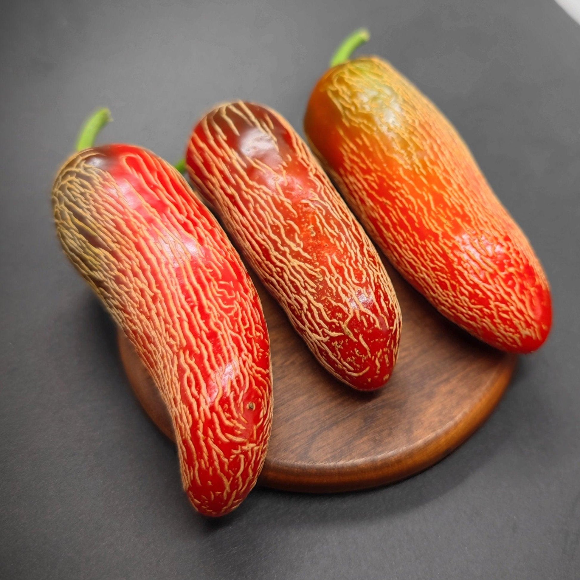 Three ripe Early Jalapenos from PepperMerchant.net with red and orange hues and wrinkled, streaked skin sit on a round wooden board against a dark background. The elongated vegetables have green stems and exhibit unique surface textures, hinting at their high Scoville rating.