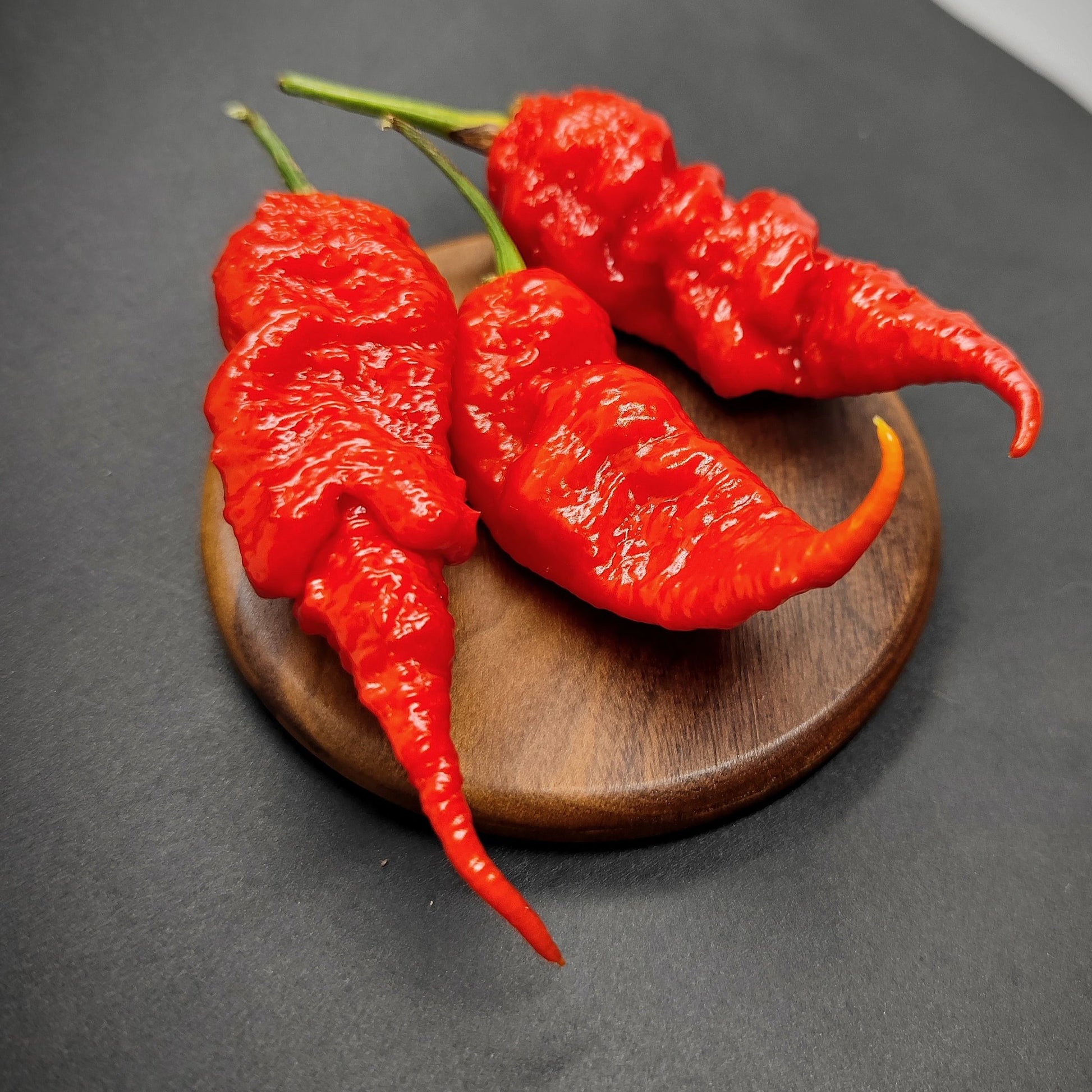 Three vibrant red Tiberius Mauler chili peppers from PepperMerchant.net, with a wrinkled surface, are displayed on a round wooden board against a dark background.