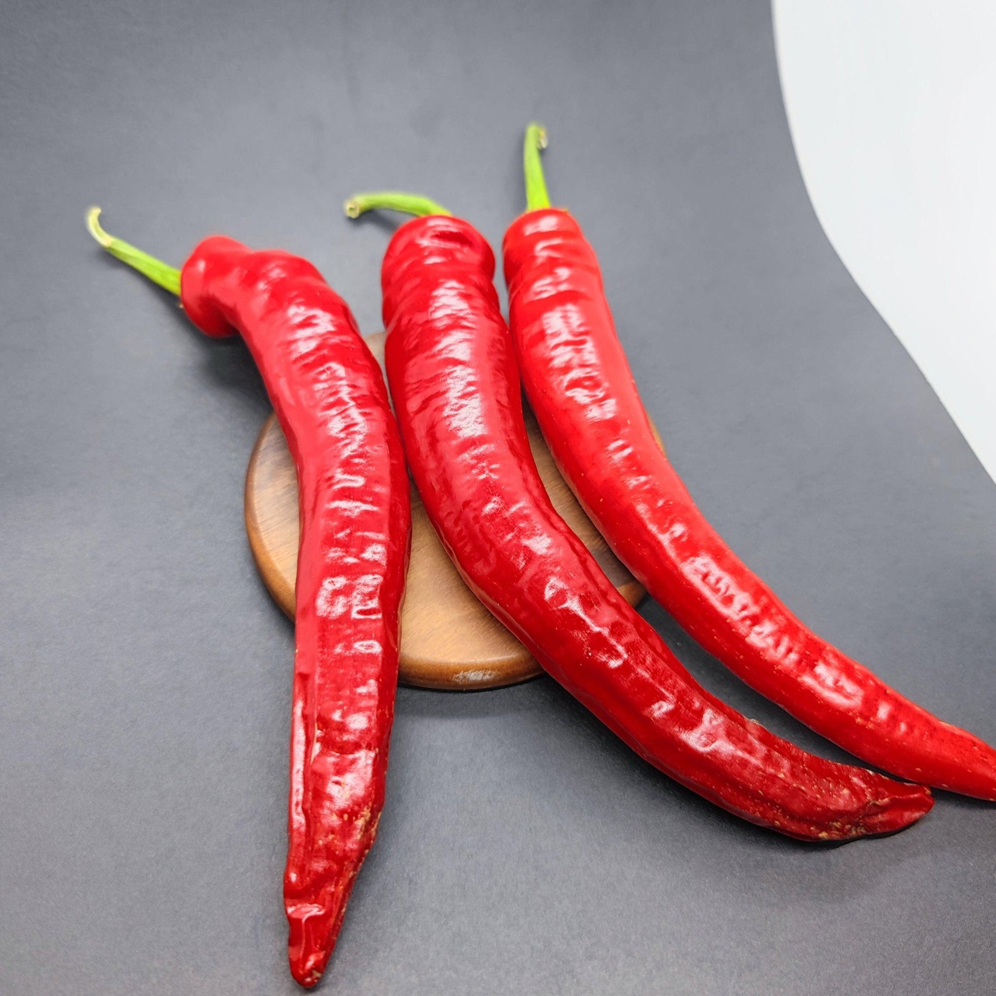 Three large cayenne peppers from PepperMerchant.net, with their glossy red appearance, sit on a small wooden round platform against a black background, suggesting their fiery potential measured in Scoville Heat Units.