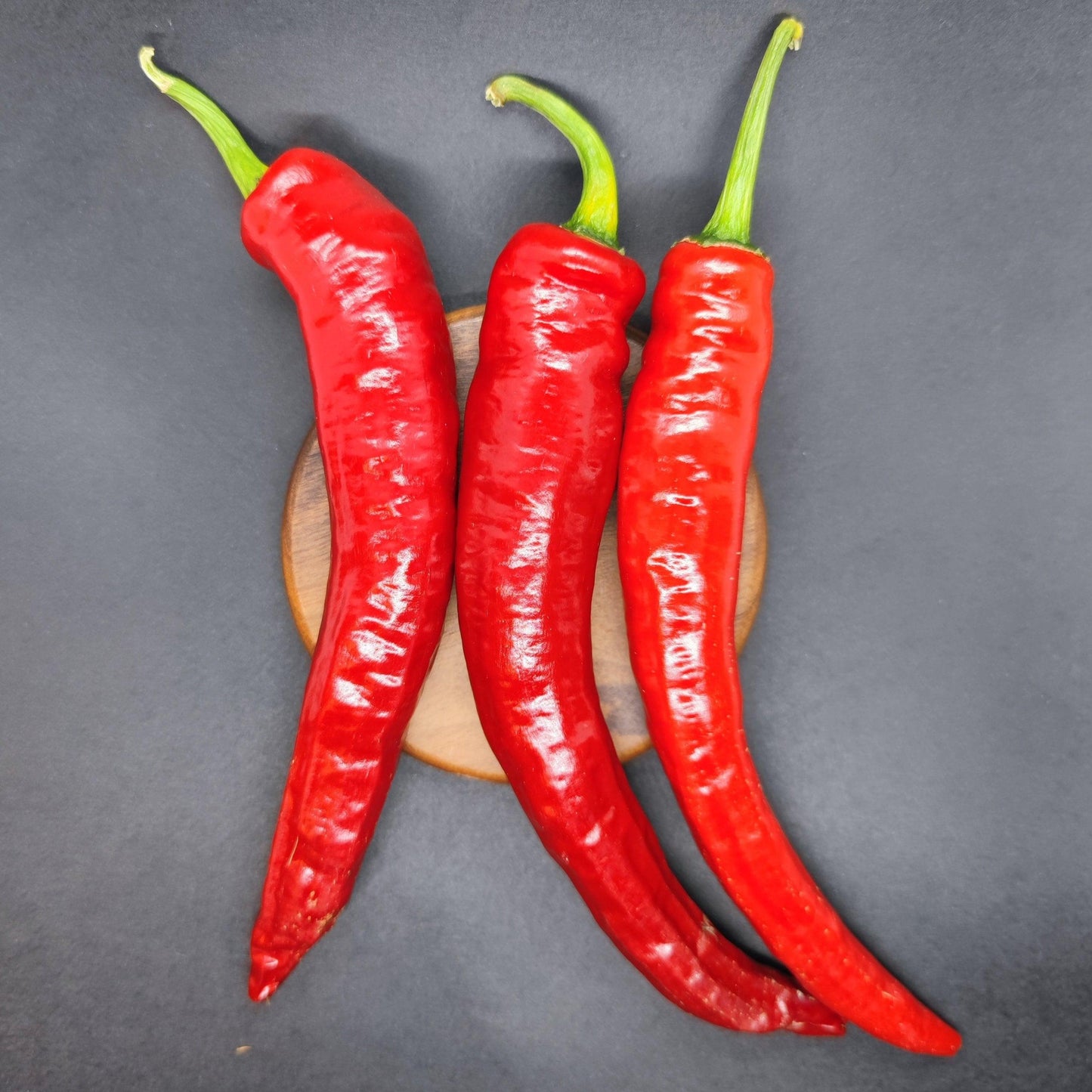 Three large Cayenne Peppers from PepperMerchant.net, vibrant red with green stems, rest on a small round wooden board against a dark background, suggesting the fiery promise of hot sauce in every bite.