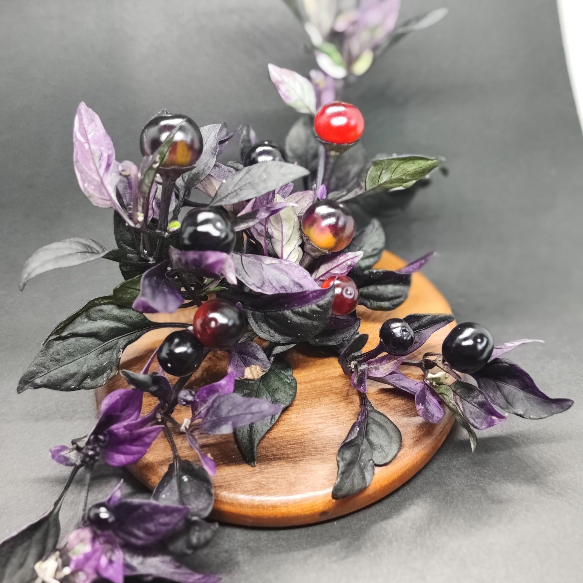 The Daybreaker by PepperMerchant.net features a branch with purple and green leaves plus glossy red, black, and dark purple berries—striped like pepper pods—displayed on a round wooden board against a dark background.