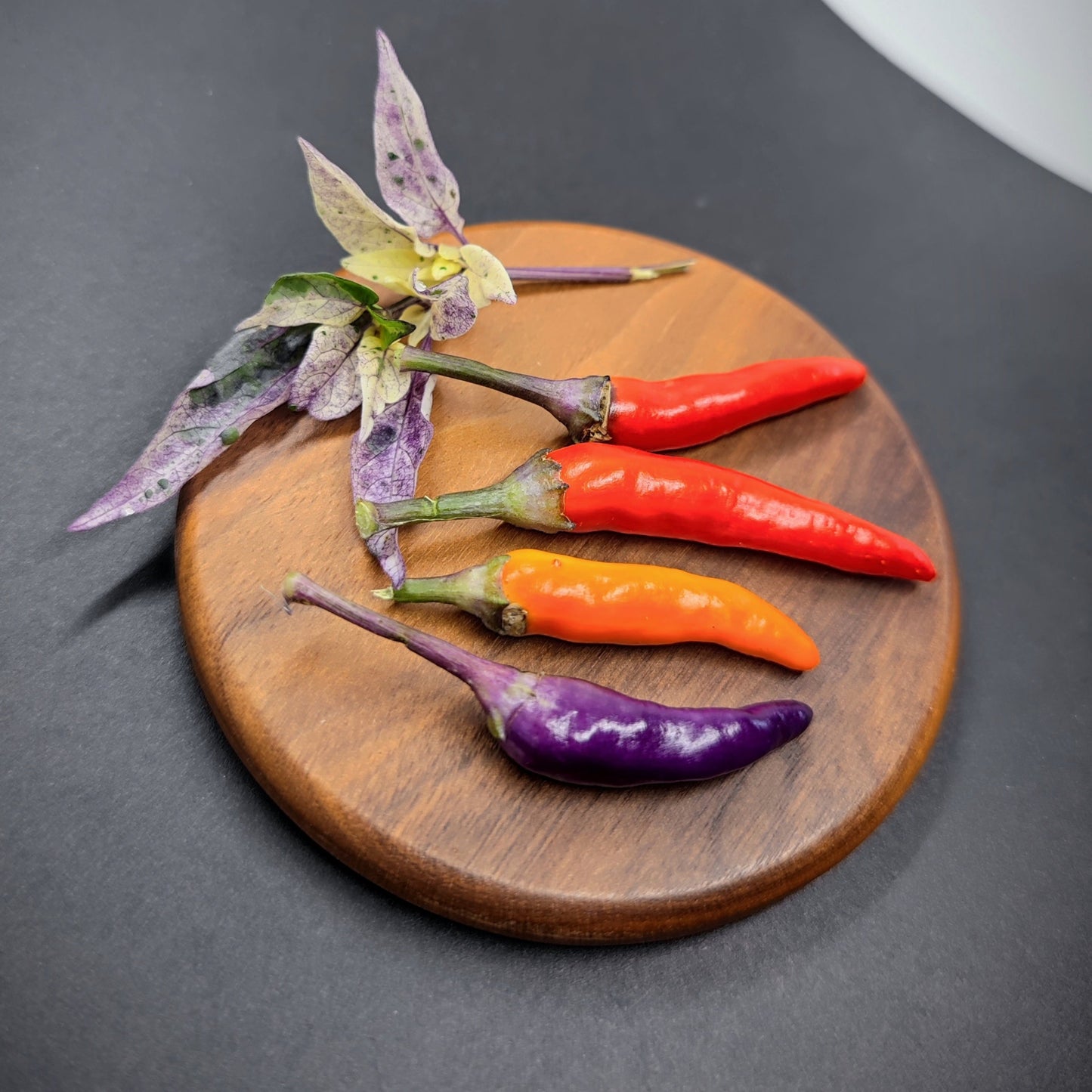 Four chili peppers—including the Purple Variegated Thai from PepperMerchant.net—are arranged on a round wooden board with vibrant leaves, set against a dark background. Ideal inspiration for gardeners seeking colorful or mild Thai chili seeds.