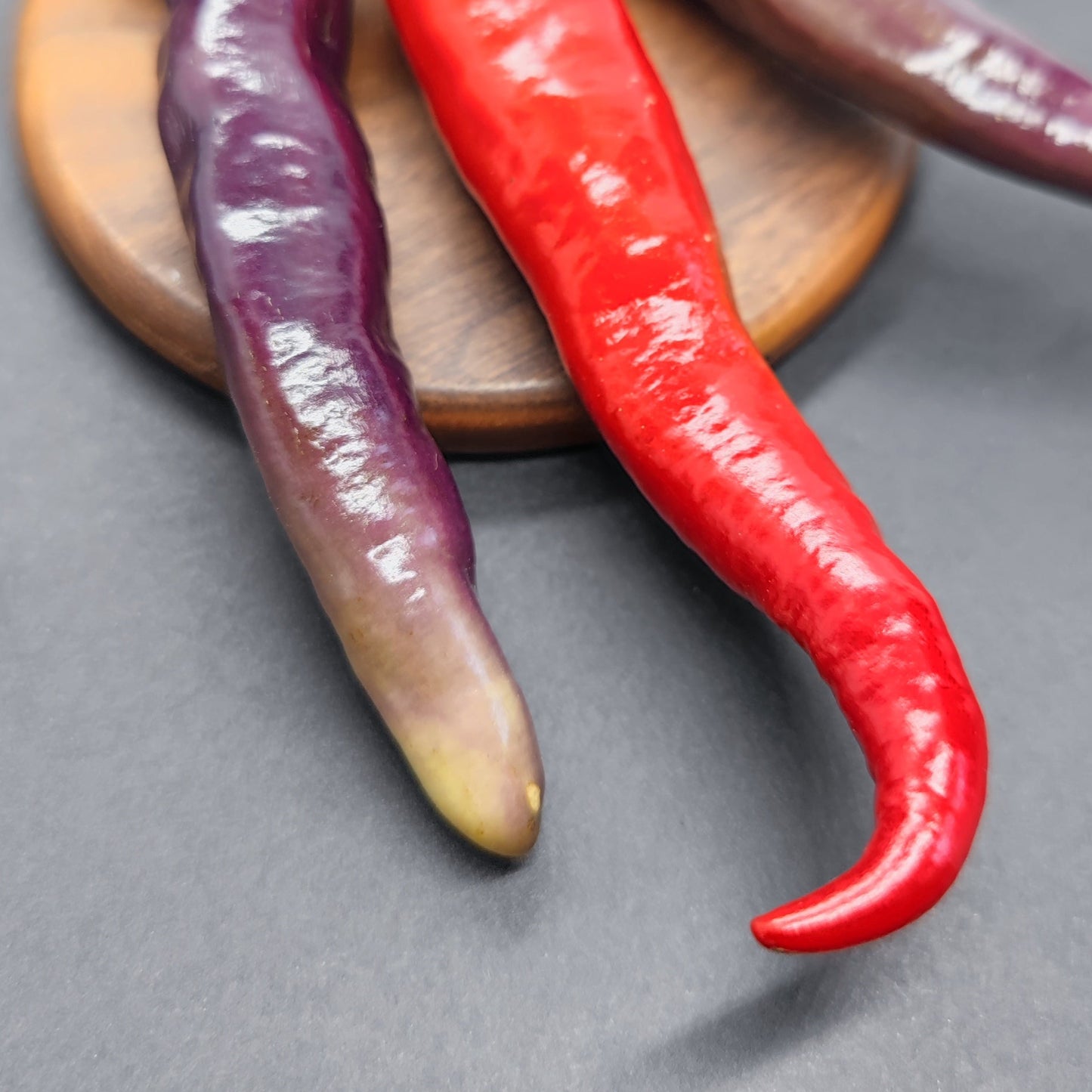 Close-up of two long chili peppers, one purple and one red, resting on a round wooden board—these may be Tiger-less Jalapeno from PepperMerchant.net, noted for their striking colors and unique hybrid characteristics.