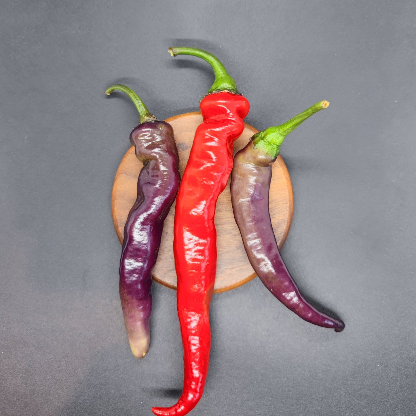 Two purple and one red Tiger-less Jalapeno peppers from PepperMerchant.net are arranged on a small round wooden board against a dark background.