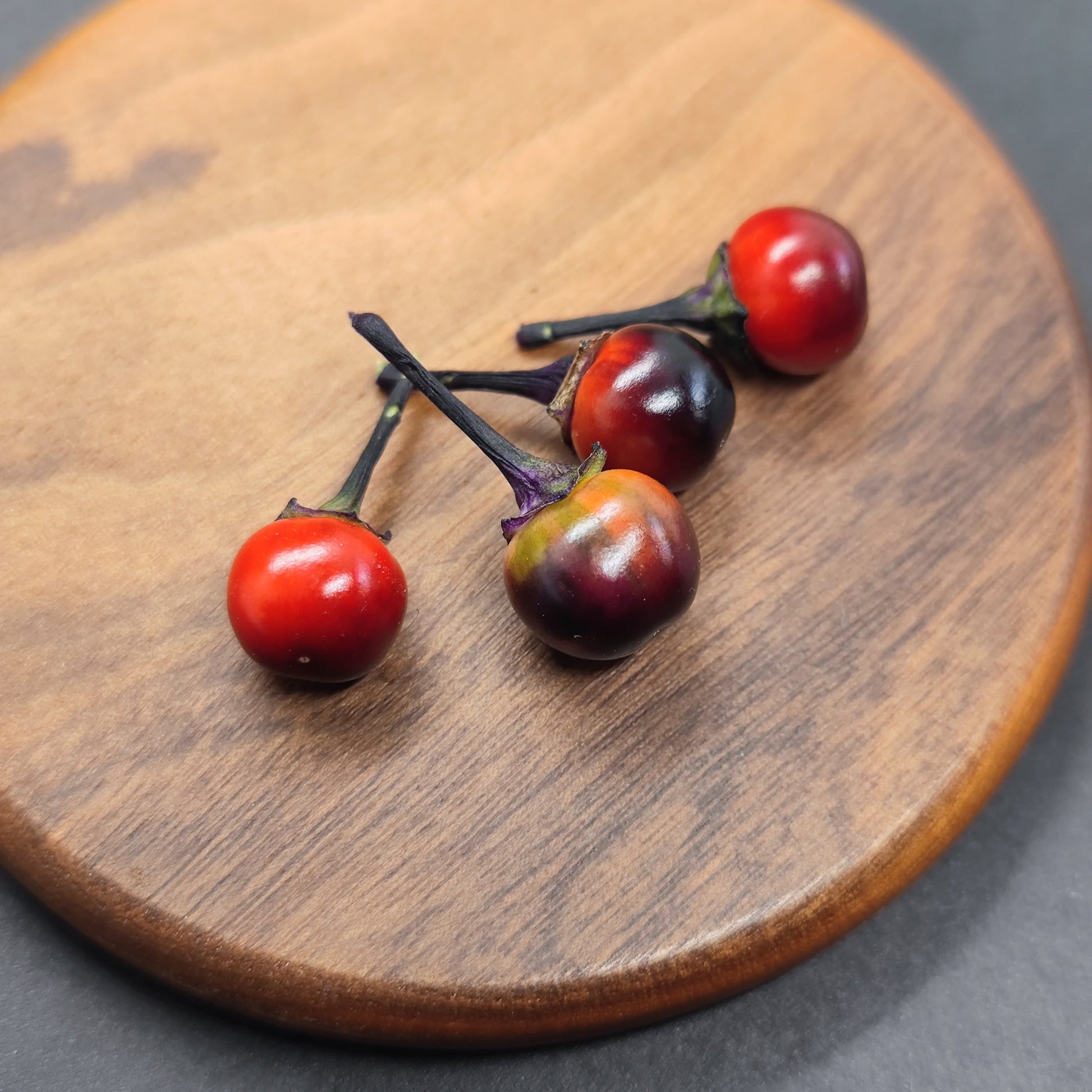 Four small, round Daybreaker peppers from PepperMerchant.net, featuring distinctive dark purple and red stripes, rest on a wooden plate against a dark background, highlighting their unique appearance.