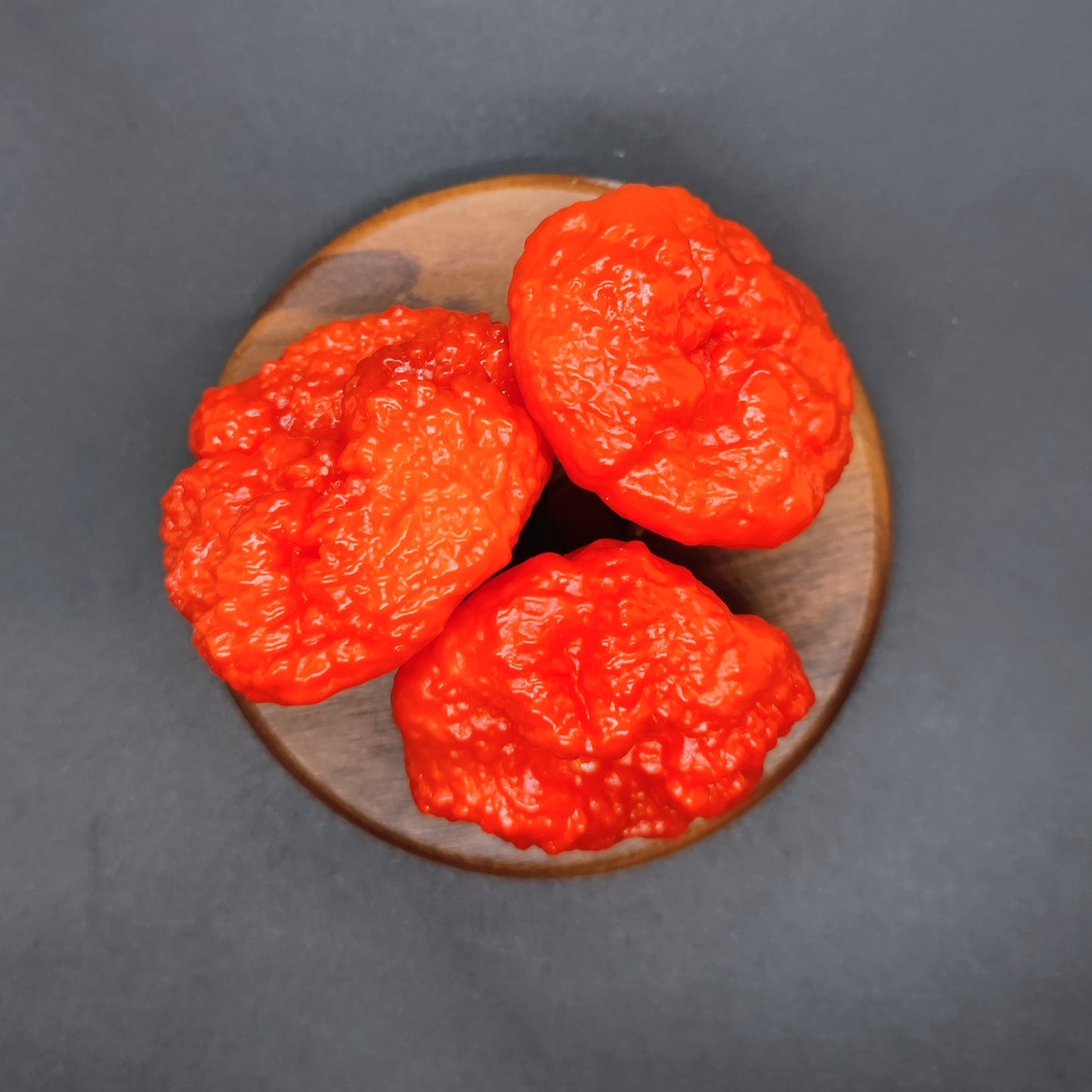 Three vivid red Scotch Brains peppers from PepperMerchant.net are arranged on a small round wooden board against a dark background.