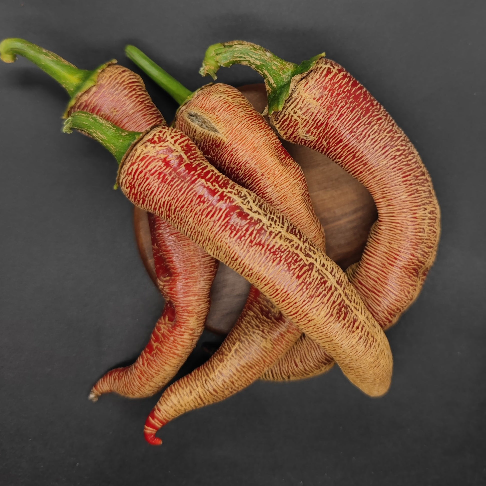 Three vibrant Vezenka peppers from PepperMerchant.net, with wrinkled skin and green stems, are displayed on a round wooden surface against a dark background, showcasing the ornamental beauty grown from Vezenka Seeds.