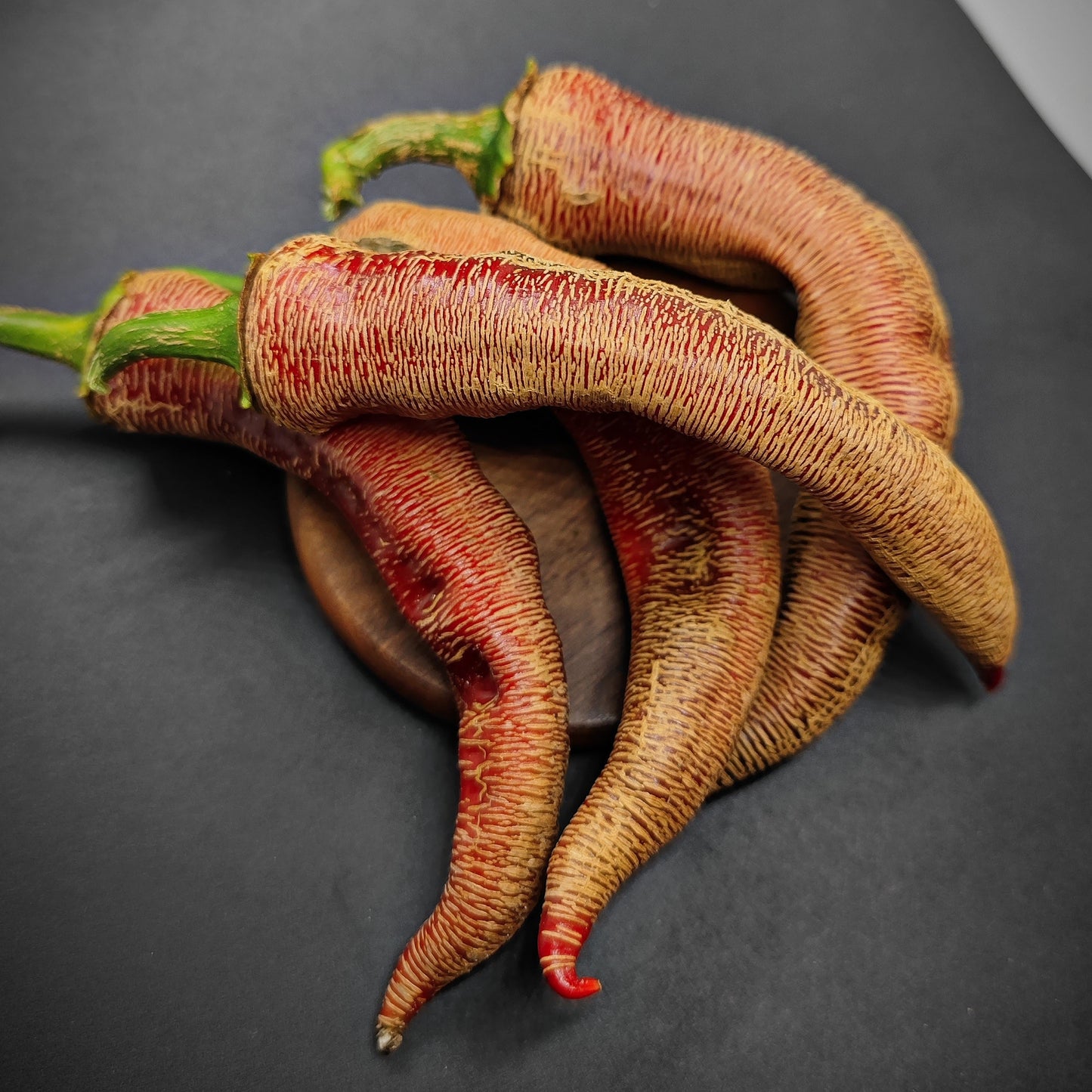 Three long, wrinkled peppers with brown, textured skin and green stems showcase the unique look of Vezenka peppers grown from Vezenka Seeds by PepperMerchant.net, arranged on a dark surface against a black background.