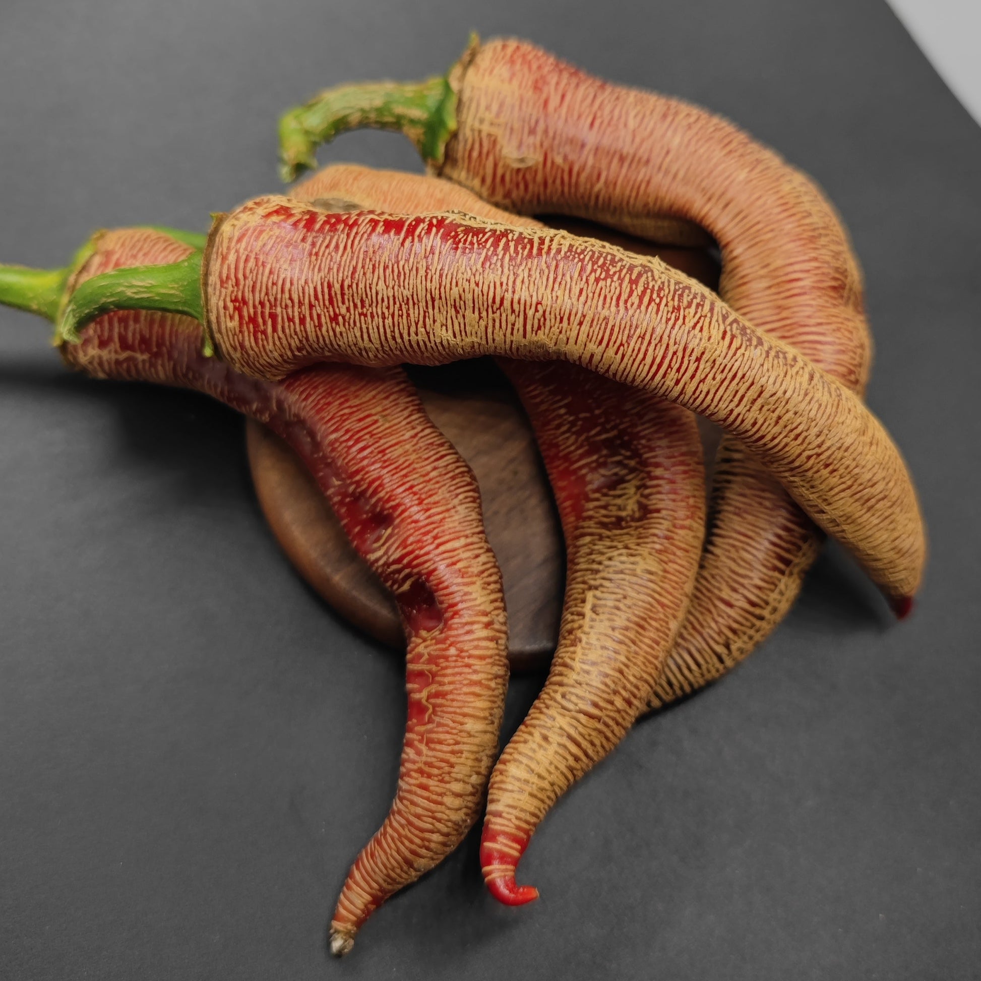 Three dried, wrinkled Vezenka peppers with green stems from PepperMerchant.net rest on a round wooden surface against a black and white background.